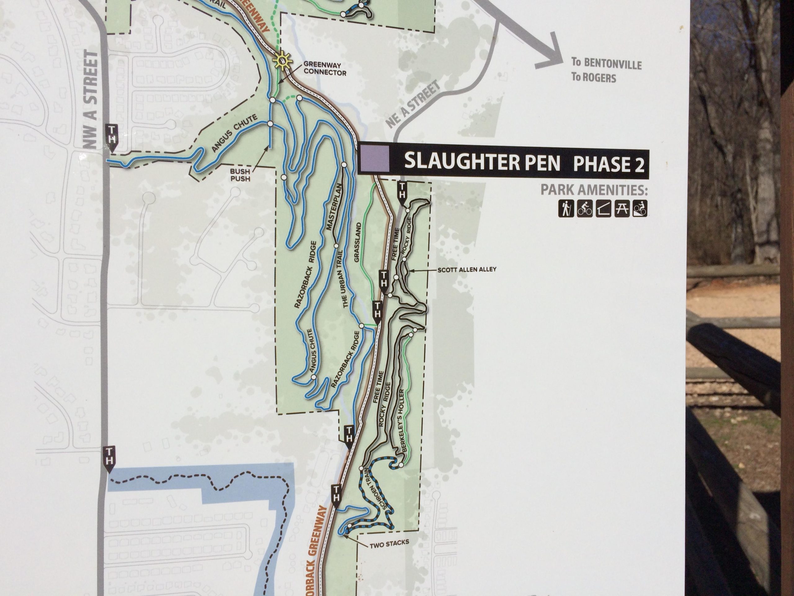 Map of Slaughter Pen Phase 2, showing trails, park amenities, and connectors to Bentonville and Rogers. Features include bike paths and walking trails, indicated by icons. Greenery and water features are illustrated along the trails. Slaughter Pen Trail mountain bike trail.