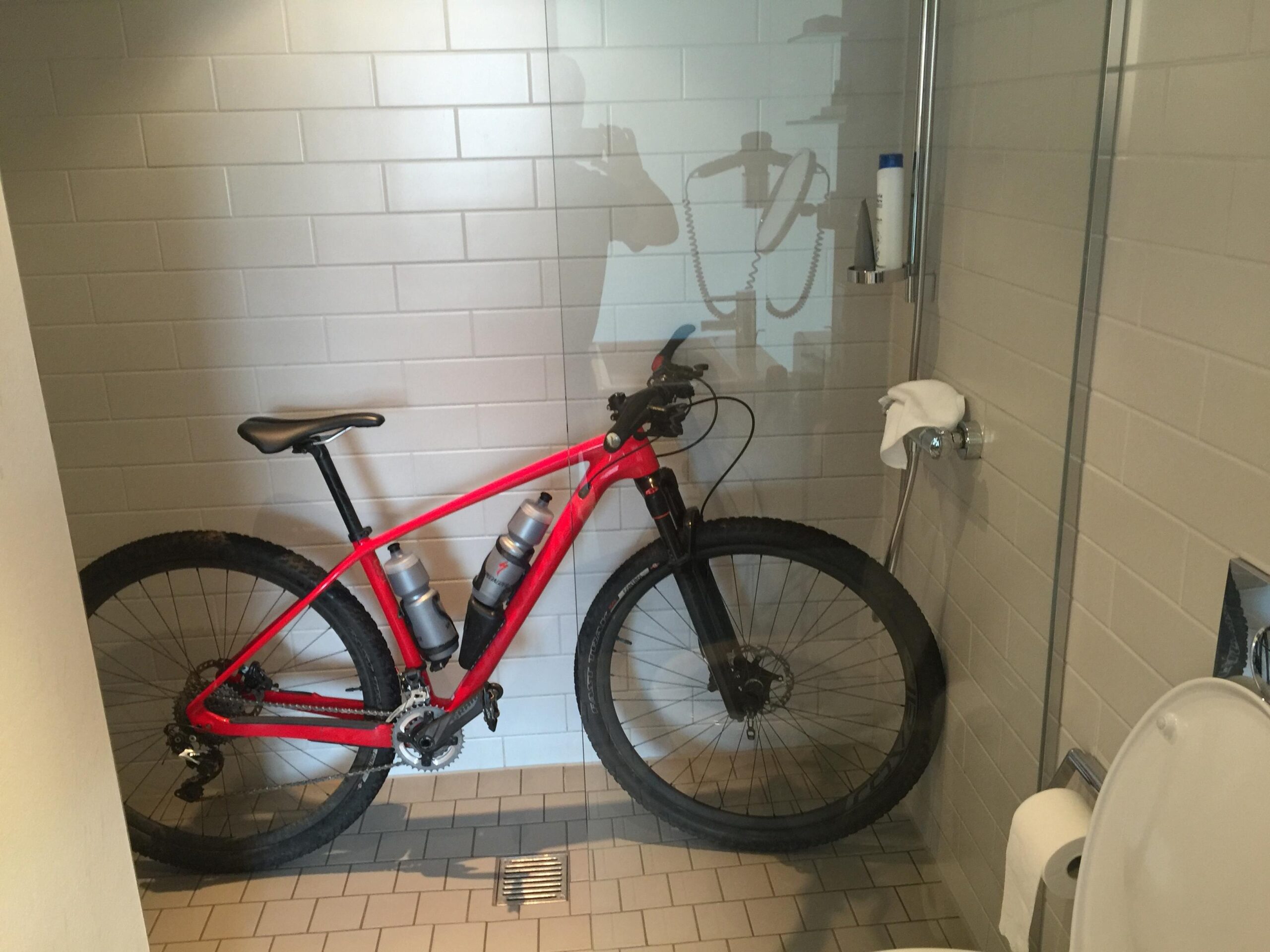 Specialized STUMPJUMPER COMP CARBON HT: A mountain bike in a shower stall, positioned next to a toilet. The bike is red with two water bottles attached, and the shower features a modern design with white tiles and chrome fixtures.