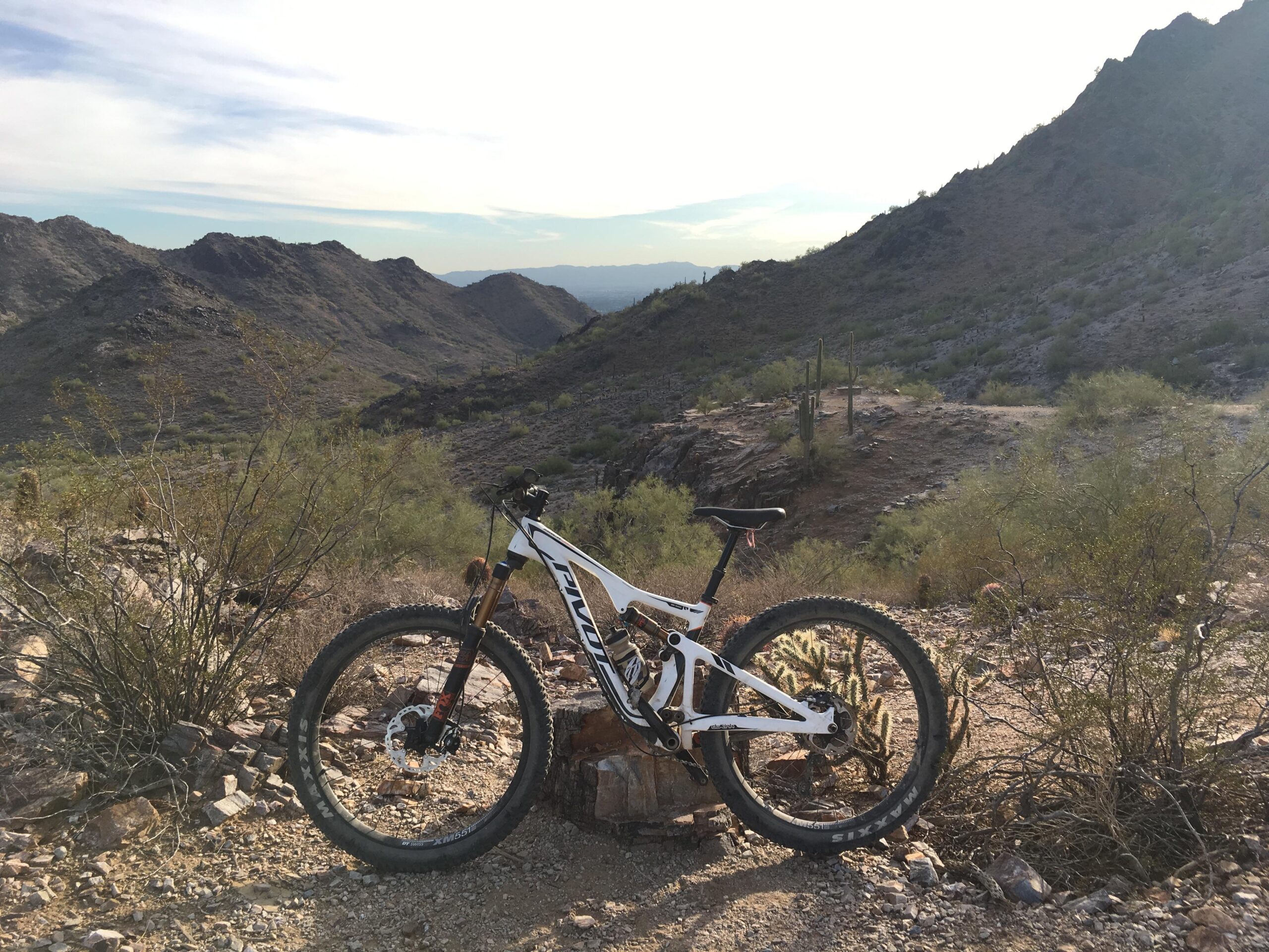 Pivot Trail 429: A mountain bike is leaning against a rock in a desert landscape, surrounded by shrubs and cacti. In the background, rugged mountains rise under a partly cloudy sky. The scene captures the beauty of outdoor biking in a natural setting.
