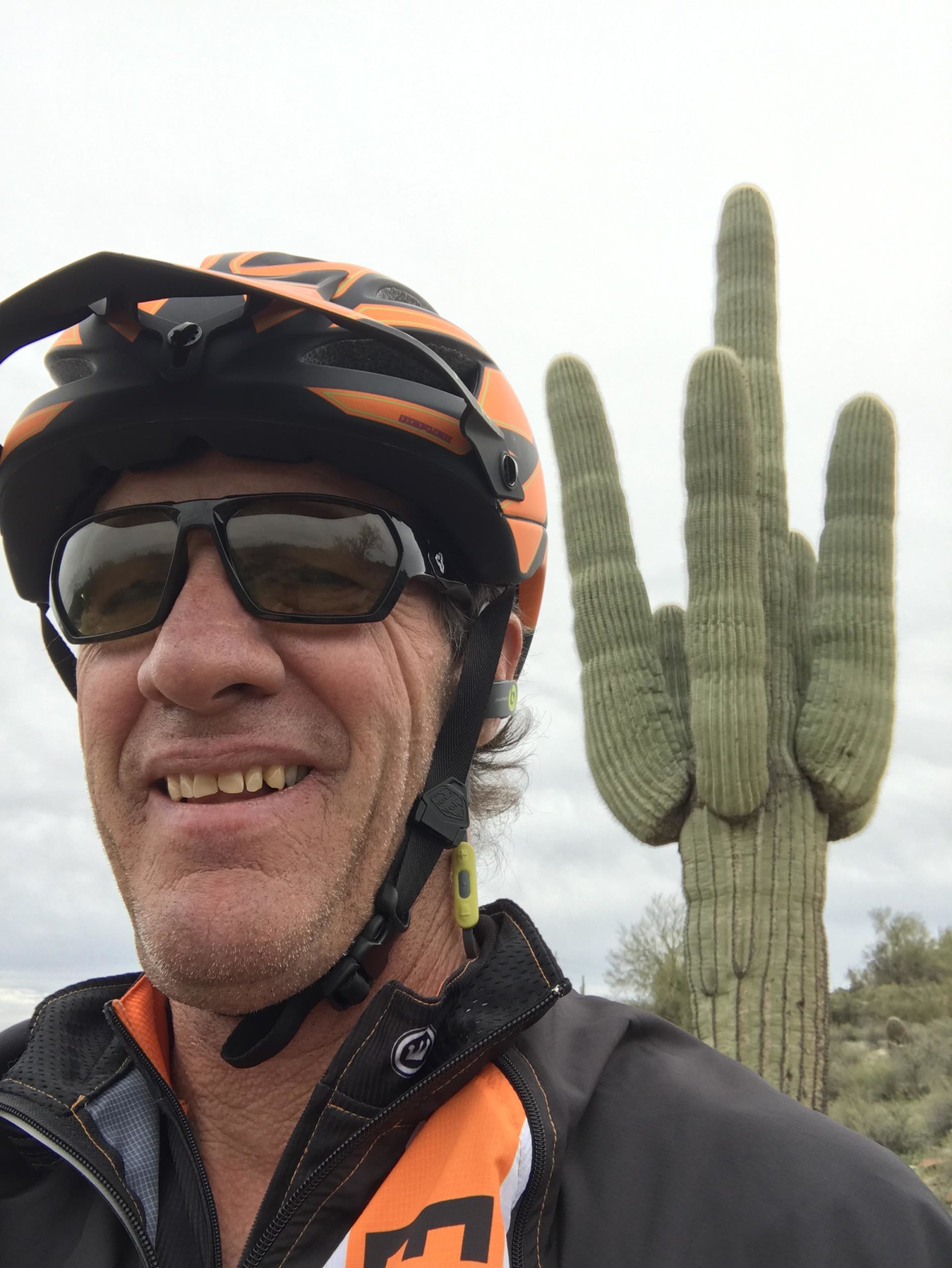 A person wearing an orange and black cycling helmet and sunglasses smiles as they take a selfie in front of a tall saguaro cactus with multiple "arms" reaching upwards. The scene is outdoors, with a cloudy sky in the background. Pemberton Loop mountain bike trail.
