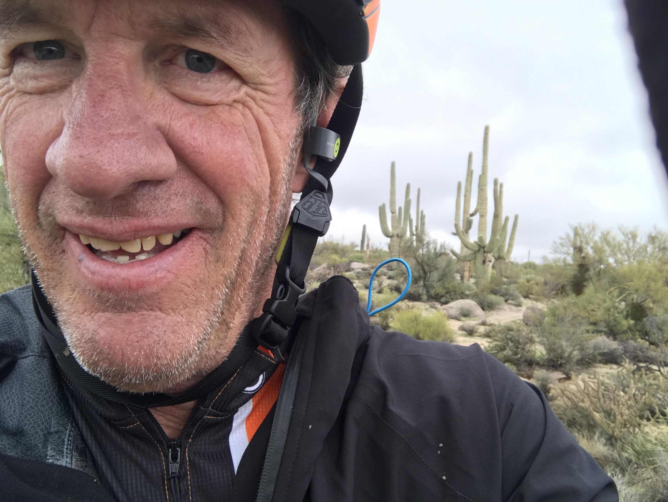 A close-up selfie of a smiling man wearing a cycling helmet and black jacket, with a desert landscape featuring tall cacti and greenery in the background under a cloudy sky. Brown's Ranch to Granite Mountain mountain bike trail.