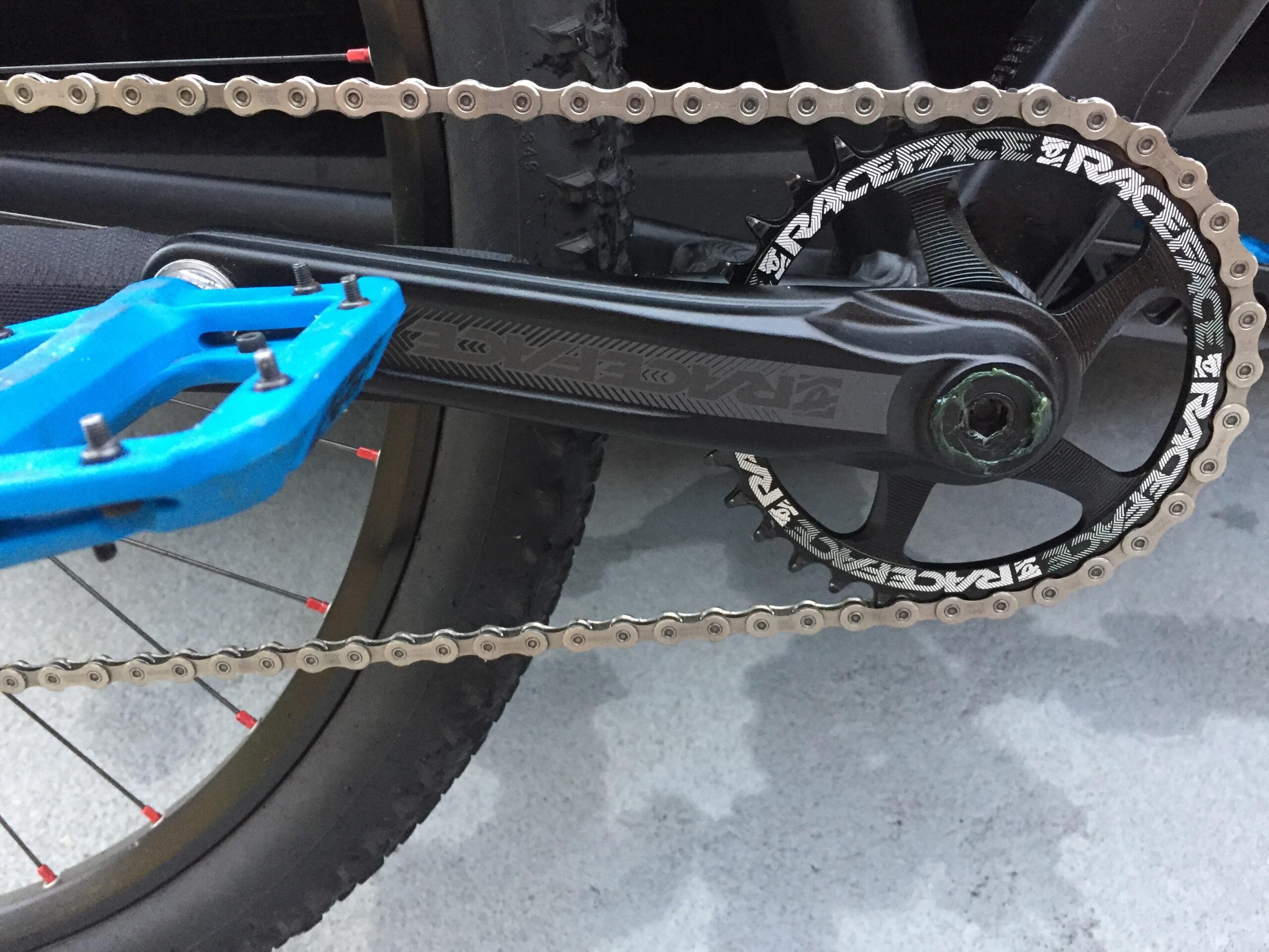 Giant Talon 29er: Close-up of a bicycle crankset with a blue pedal attached. The crank arm and chainring are marked with the "Raceface" logo, and a silver chain runs along the chainring. A portion of the bike’s tire is visible in the background, with spokes featuring red accents. The surface beneath the bike is gray.