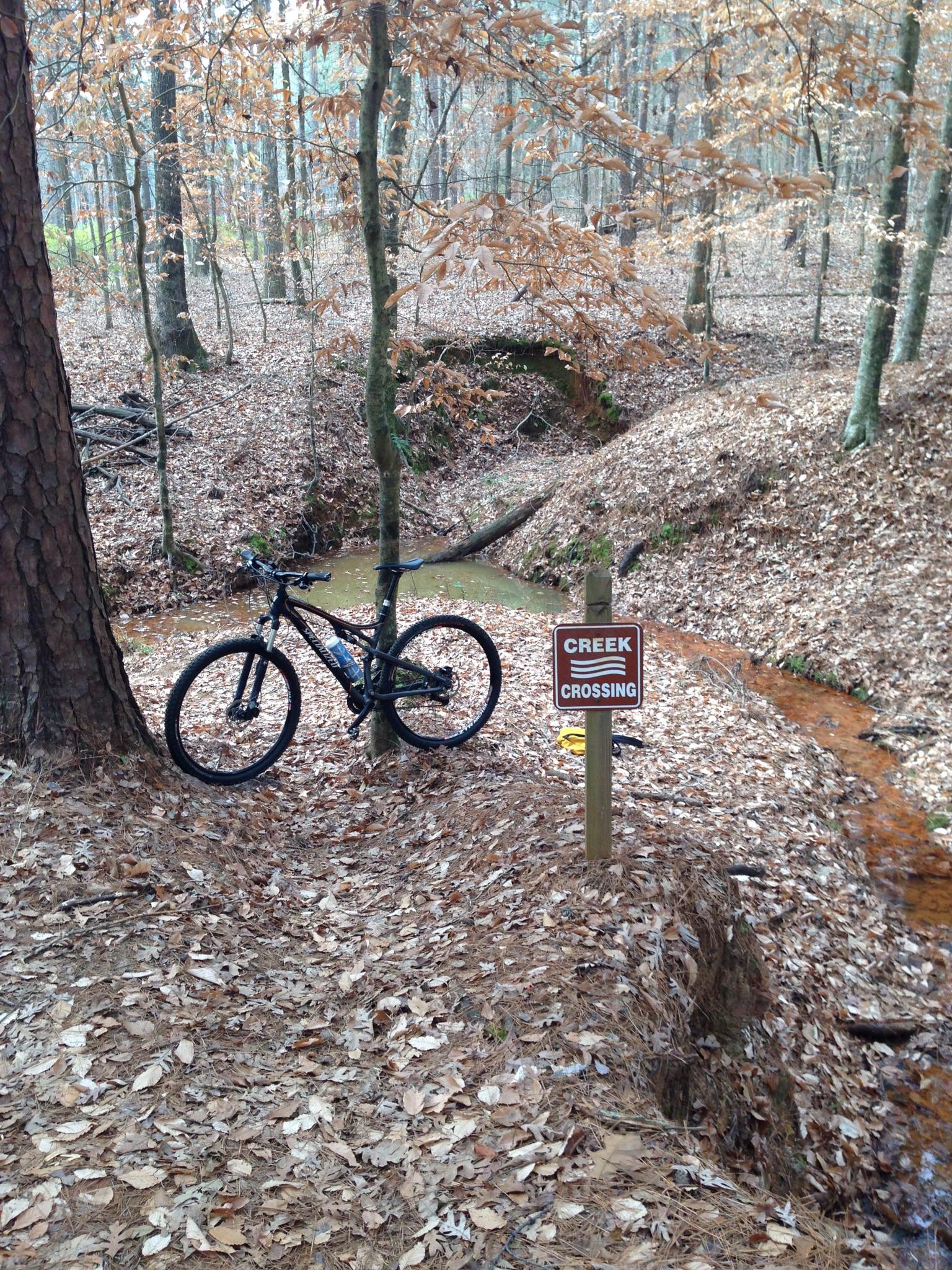 A mountain bike is parked next to a creek crossing sign in a wooded area. The ground is covered with fallen leaves, and trees with sparse foliage are visible in the background, creating a serene autumn atmosphere. A shallow creek flows nearby, surrounded by earthy banks. Lincoln Parish Park mountain bike trail.