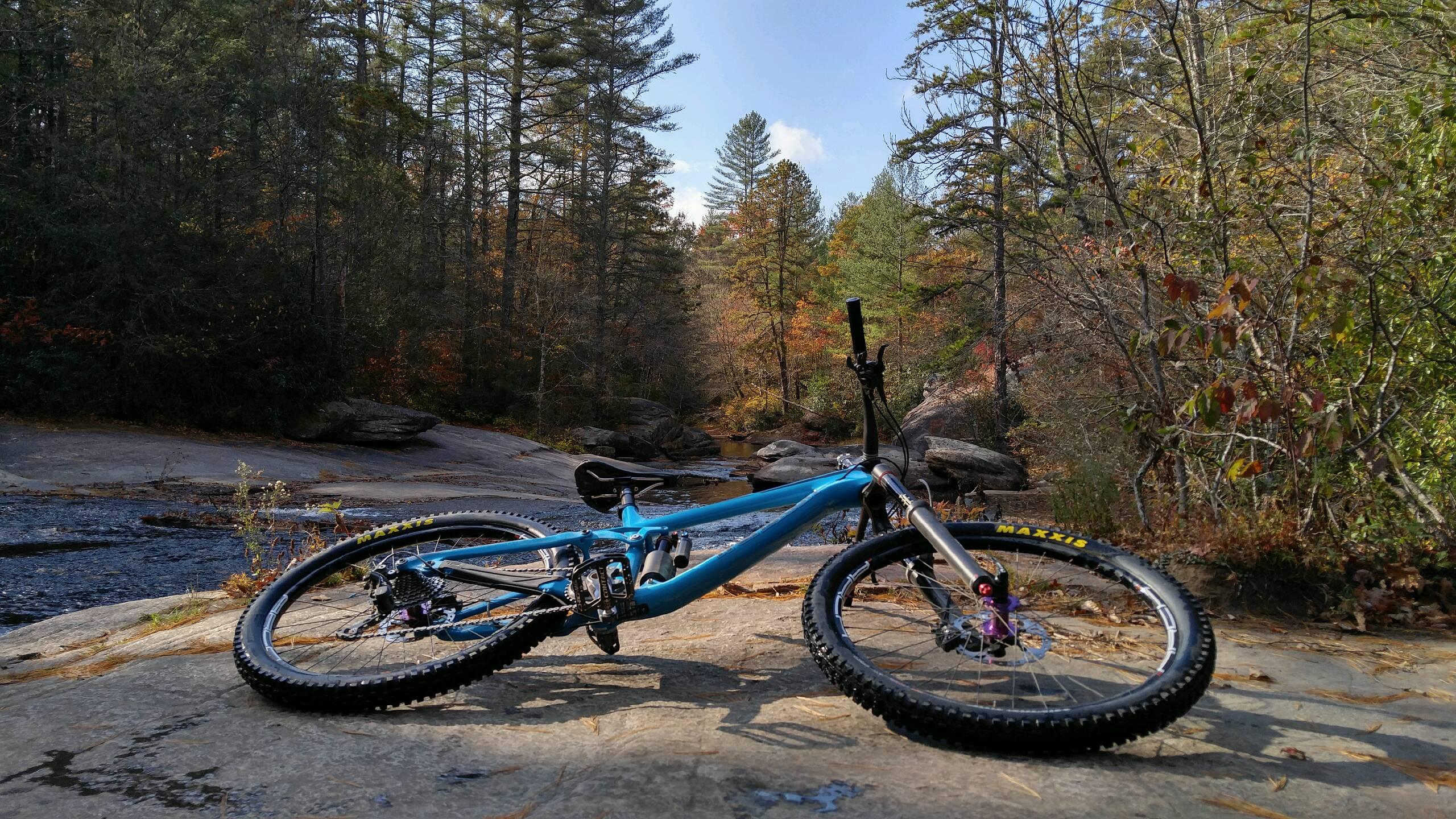 Transition Patrol: A blue mountain bike lying on a rock by a stream, surrounded by trees displaying autumn foliage. The background features a peaceful landscape with a partially cloudy sky and hints of colorful leaves.