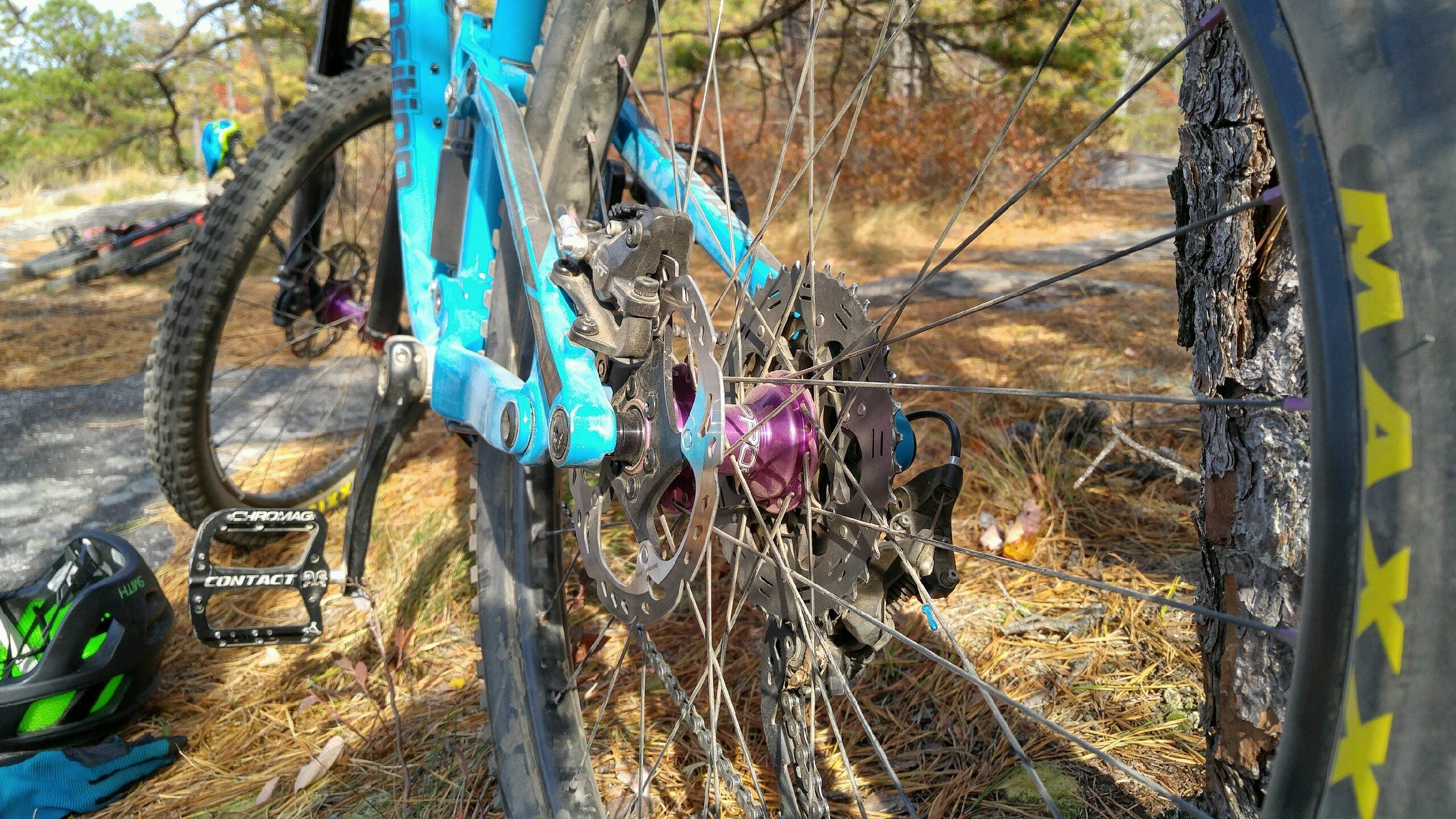 Transition Patrol: Close-up view of a mountain bike's rear wheel, showcasing the purple hub, disc brake, and gear mechanism. In the background, a blurred second bike is visible along with some fallen pine needles on the ground. A black pedal with "Chromag Contact" branding and a green and black helmet are placed nearby.