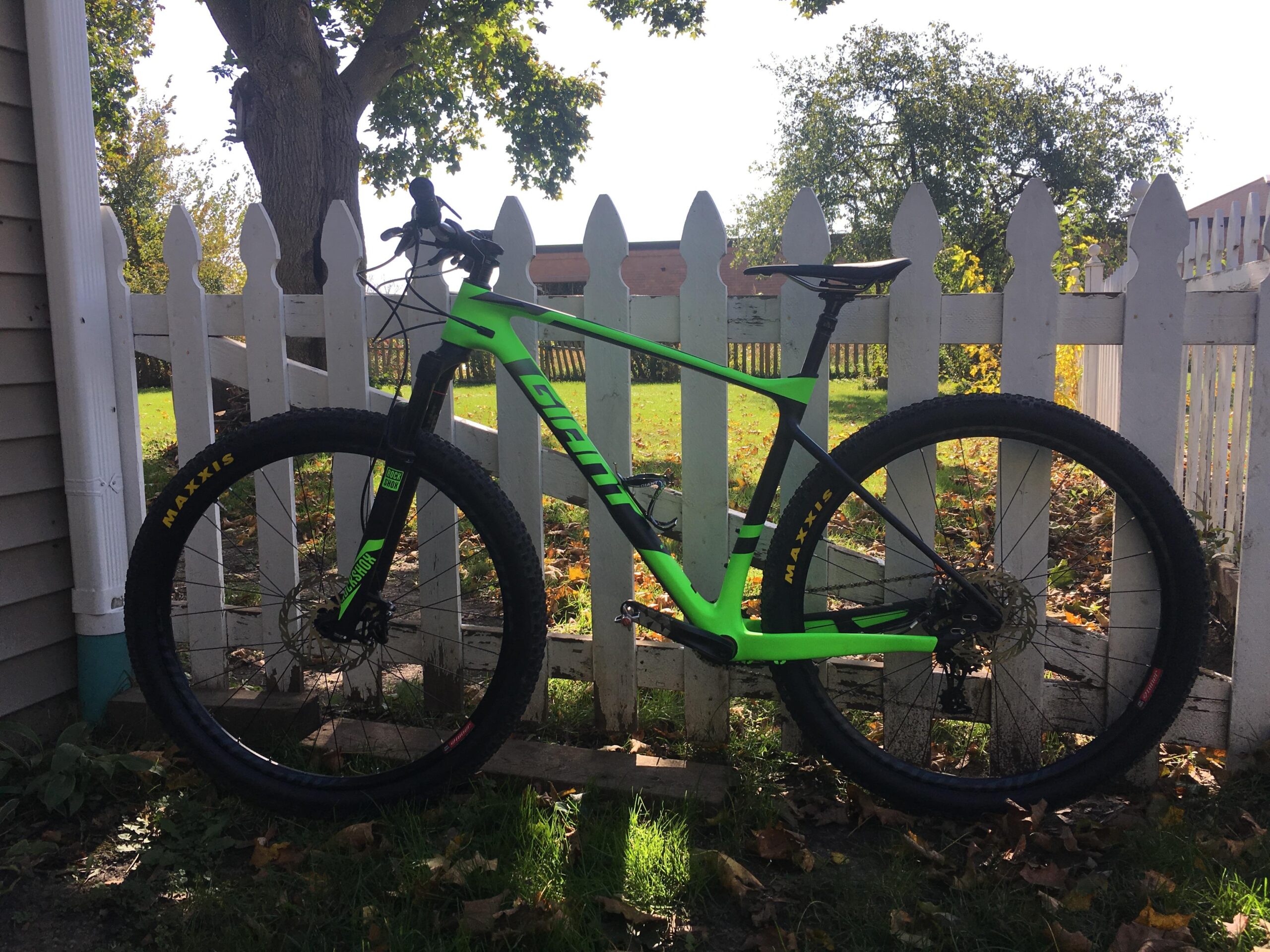 Giant XTC 27.5 2: A vibrant green and black mountain bike leaning against a white picket fence, with lush green grass and scattered autumn leaves in the background.