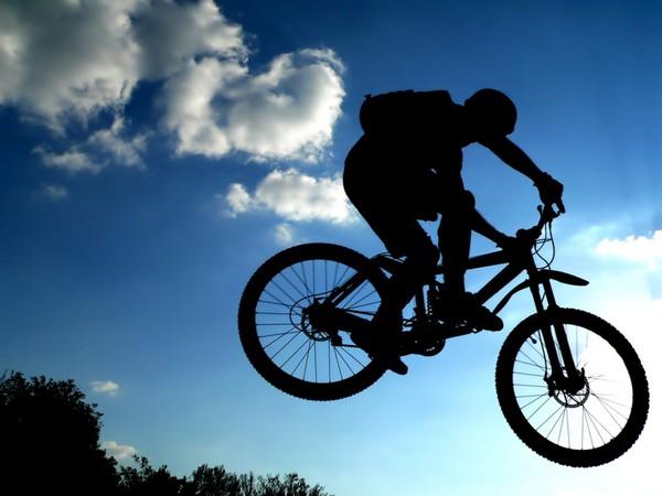 A silhouette of a mountain biker performing a jump against a backdrop of blue sky and fluffy white clouds. The sun is partially visible, adding a glowing effect around the biker.