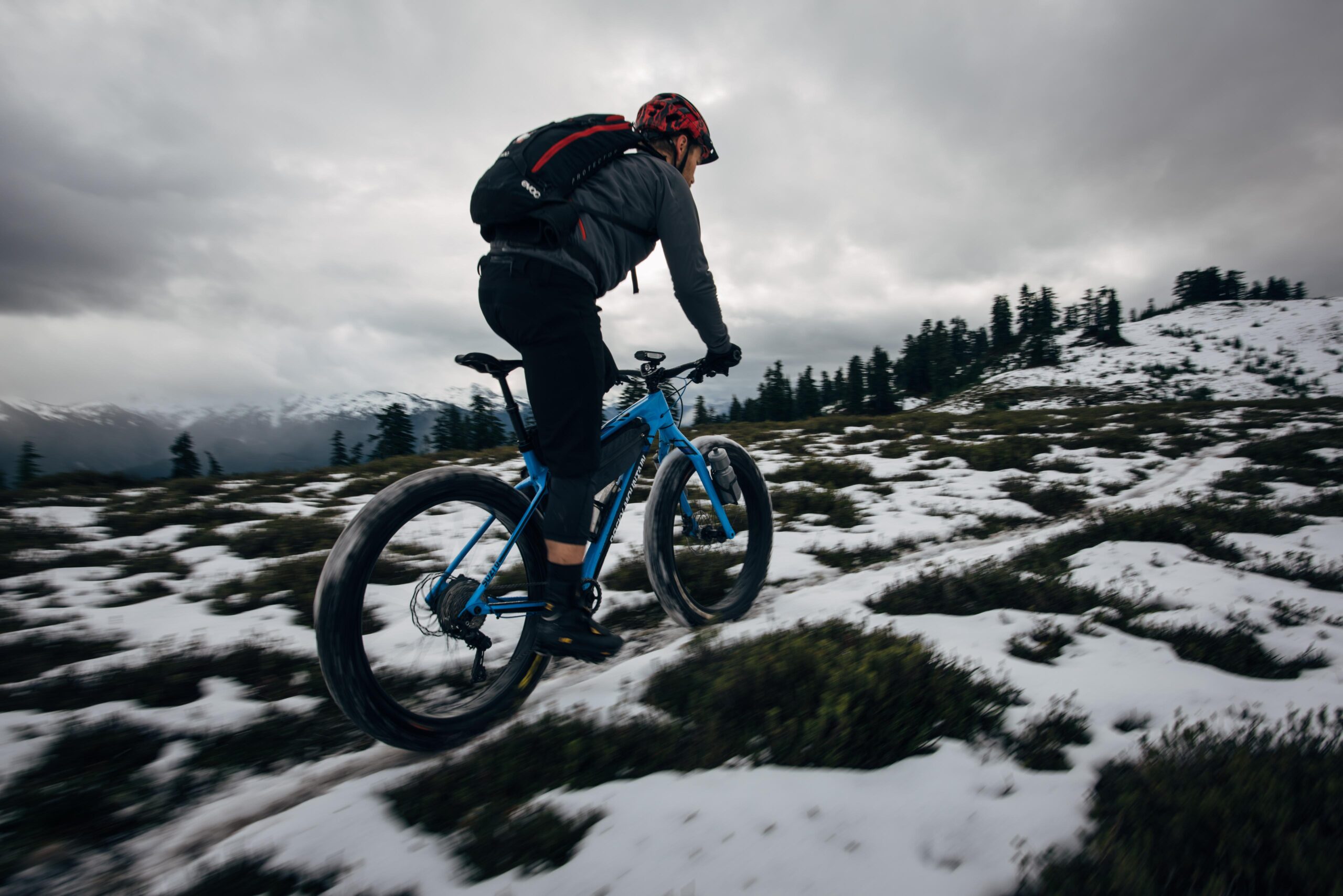 Rocky Mountain Suzi Q: A cyclist riding a blue mountain bike on a snow-covered trail in a mountainous landscape, with dark clouds overhead and pine trees in the background.