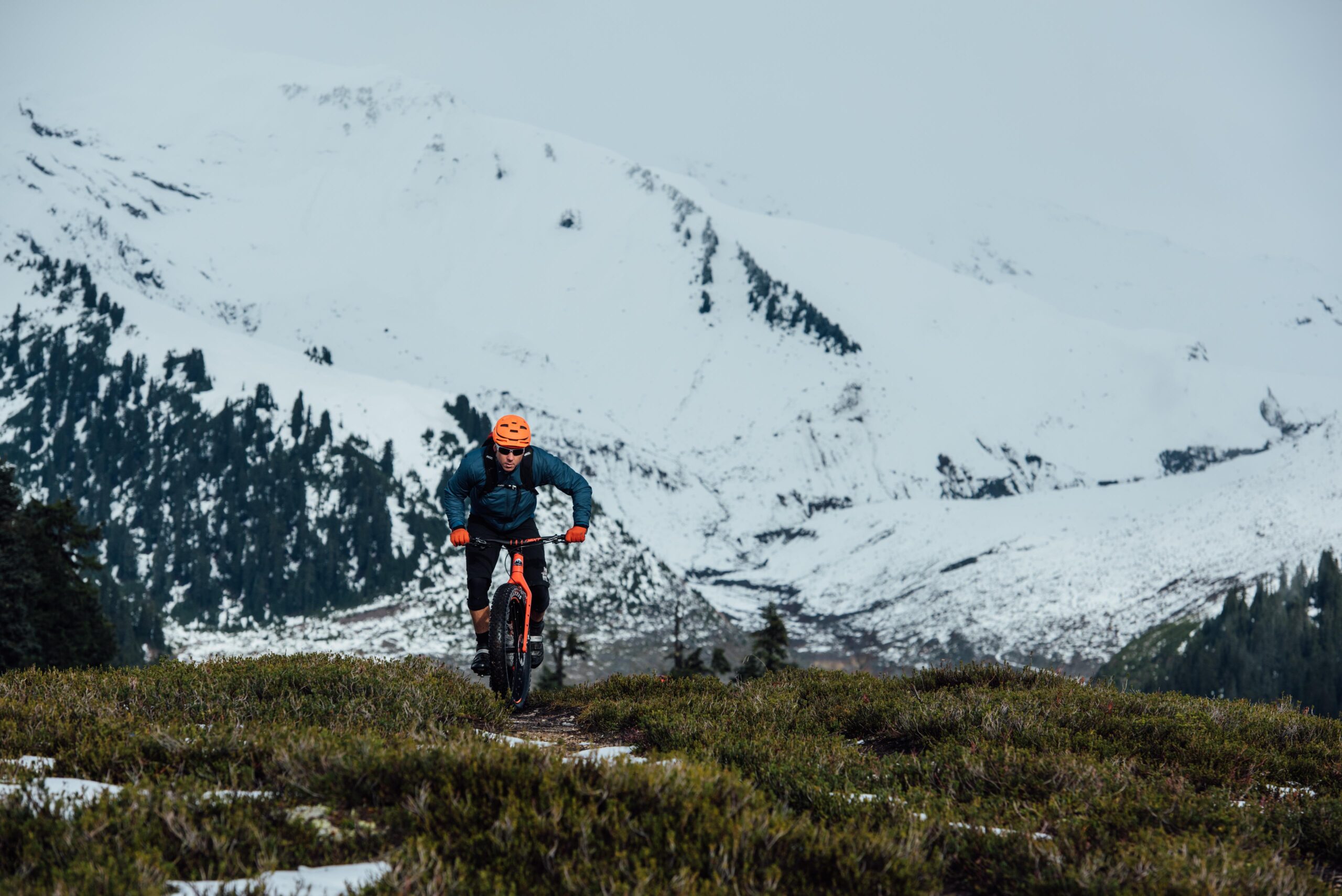 Rocky Mountain Suzi Q: A person riding a mountain bike on a grassy trail surrounded by snowy mountains. The cyclist is wearing an orange helmet and is dressed in a blue jacket and black shorts, navigating through a rugged outdoor landscape.