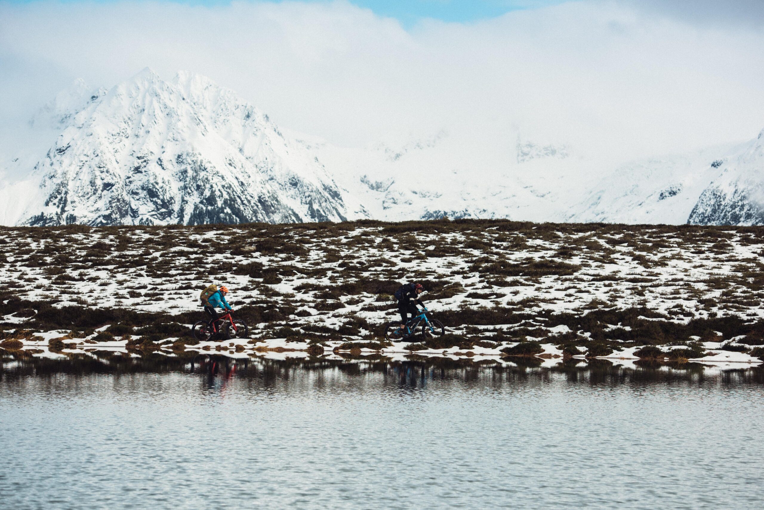 Rocky Mountain Suzi Q: Two mountain bikers ride along a snowy landscape beside a calm lake, with snow-capped mountains in the background under a partly cloudy sky. The terrain is rocky and covered with patches of snow.