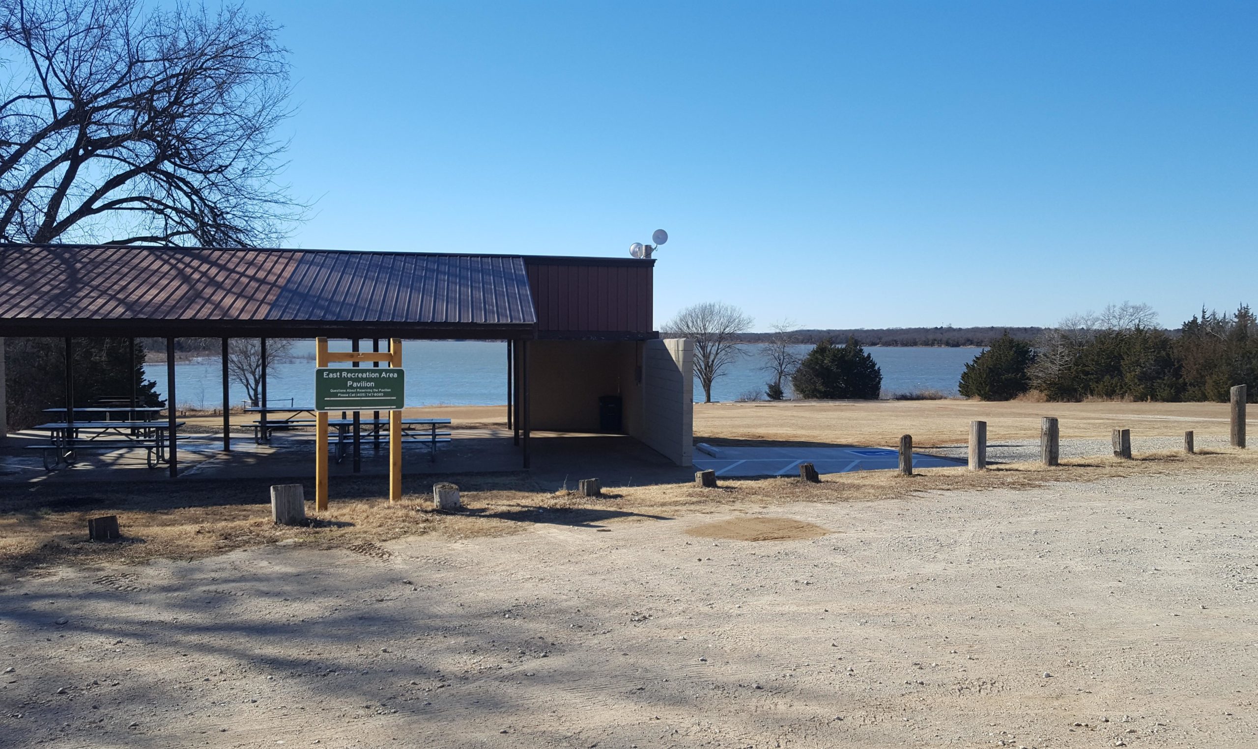 A view of a pavilion at the East Recreation Area, featuring picnic tables under a roof and a sign indicating its name. In the background, there is a scenic view of a lake and a clear blue sky. The area is surrounded by grassy land and a few trees. The ground is gravel and features wooden posts lining the area. McMurtry Trail mountain bike trail.