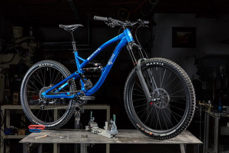 A blue mountain bike displayed on a workbench in a workshop setting, with a dark background. The bike features multiple gears, thick tires labeled "Maxxis," and a front suspension fork, highlighting its design for rugged terrain. Tools and equipment are visible in the background, emphasizing a repair or assembly environment.