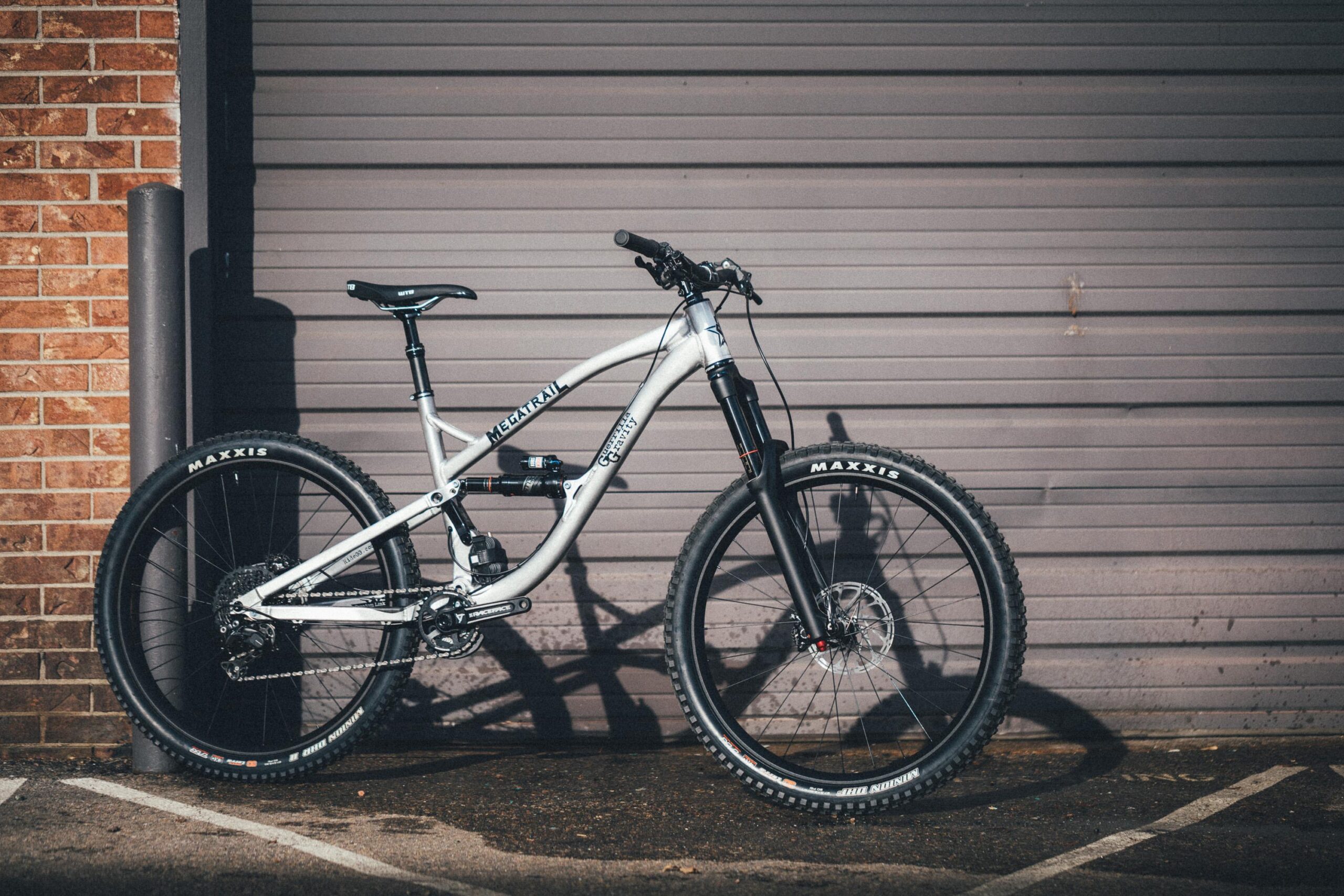Guerrilla Gravity Megatrail: A silver mountain bike with the word "Megatrail" displayed on the frame, leaning against a textured gray garage door. The bike features a front suspension fork, wide tires with "Maxxis" branding, and a sleek design suitable for off-road cycling.