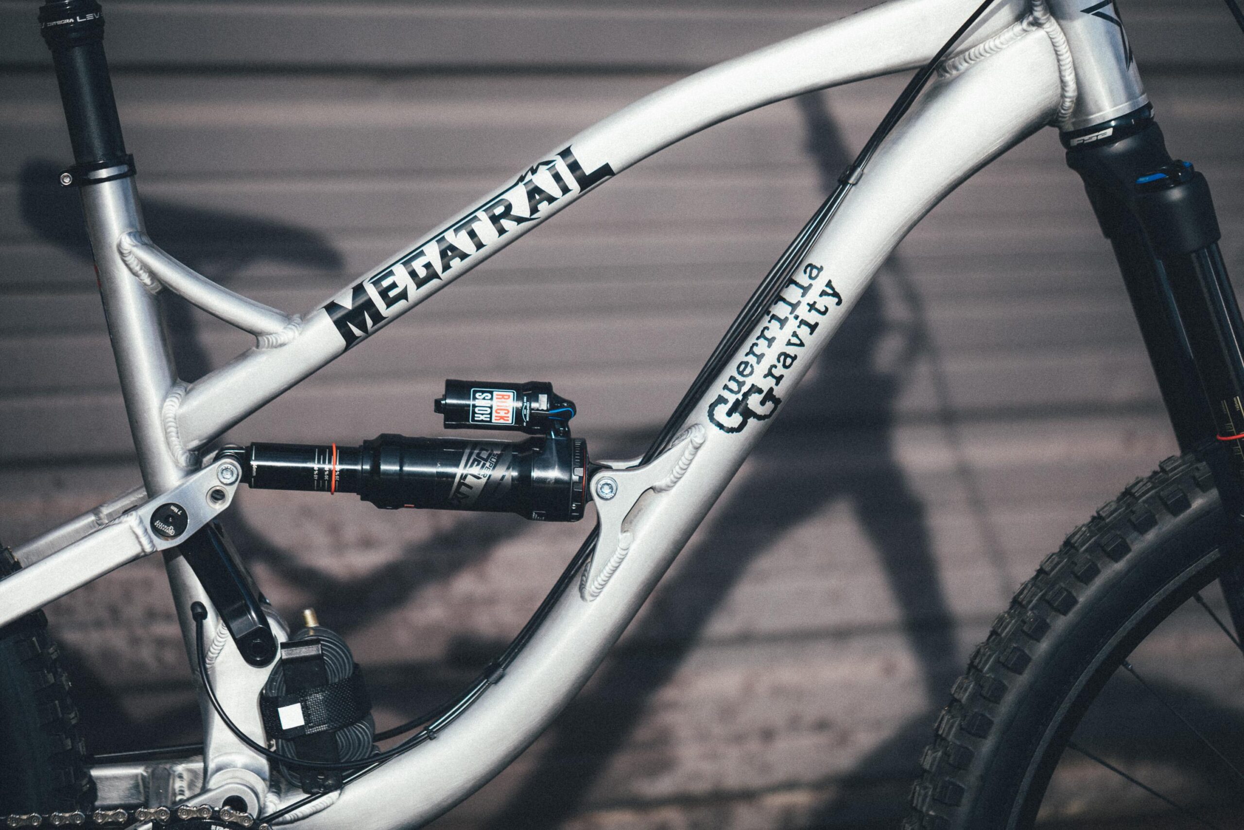 Guerrilla Gravity Megatrail: A close-up view of a mountain bike frame featuring the brand name "MEGATRAIL" prominently displayed on the top tube. The frame showcases a durable design with a suspension system, including a shock absorber labeled "RockShox." The bike's body is predominantly silver with black lettering, set against a blurred background that suggests a garage or workshop environment.