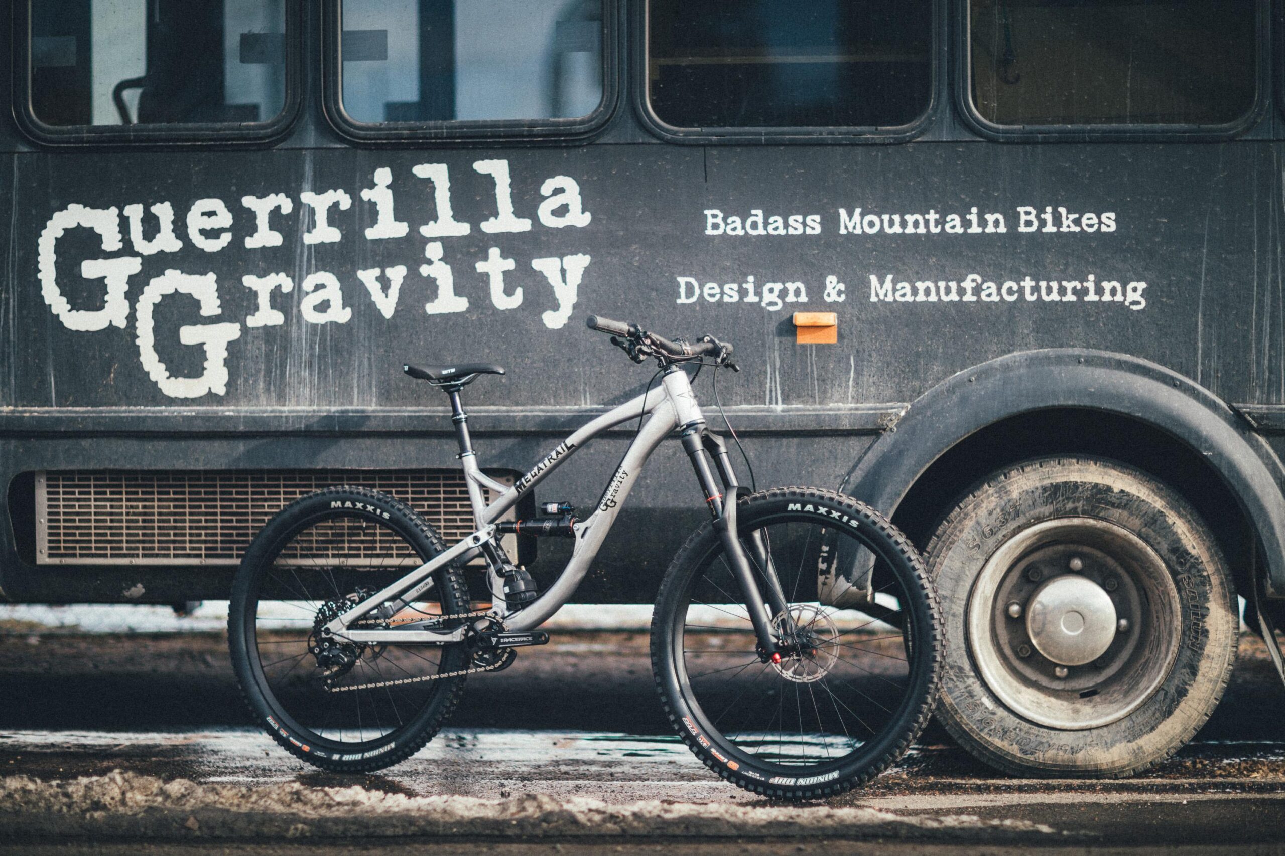 Guerrilla Gravity Megatrail: A mountain bike leaning against a rugged, vintage bus featuring the text "Guerrilla Gravity" and the phrases "Badass Mountain Bikes" and "Design & Manufacturing." The scene is set on a gravel surface with hints of mud, conveying an adventurous outdoor vibe.