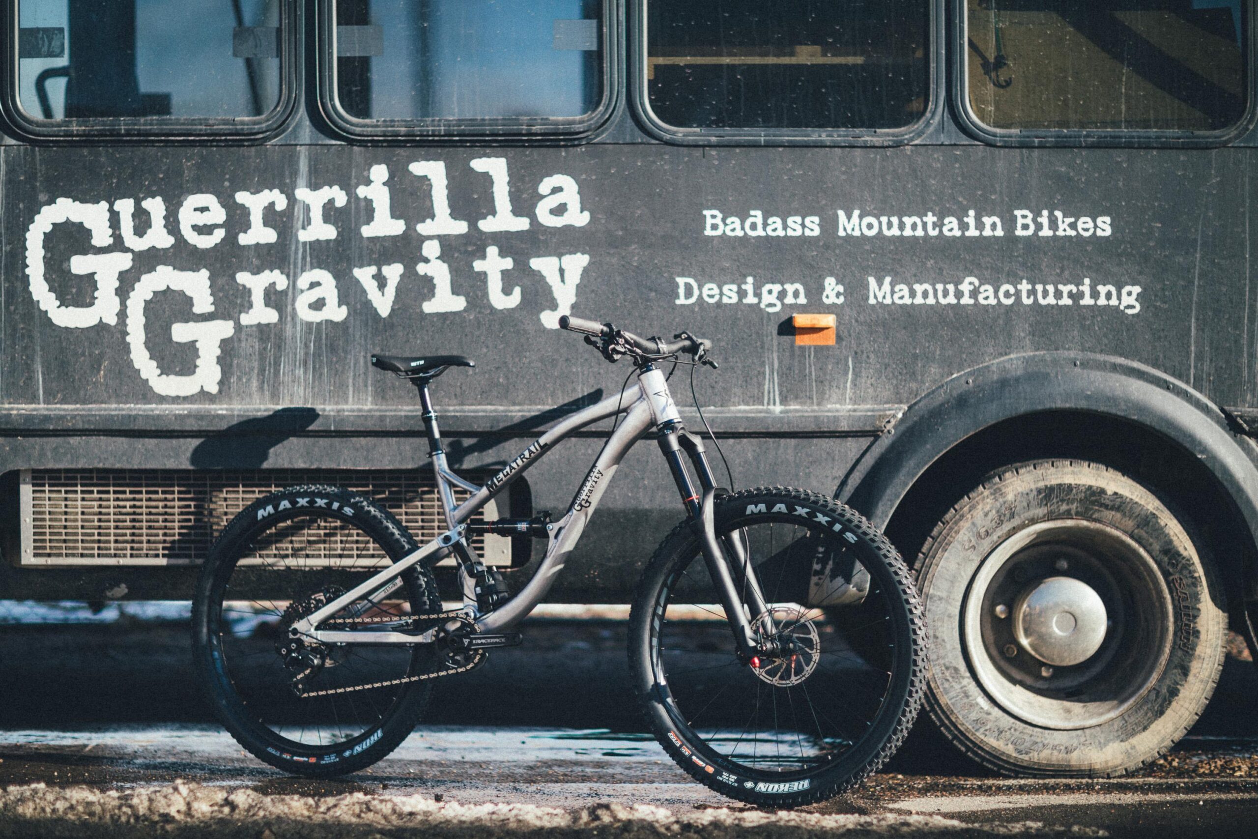 Guerrilla Gravity Megatrail: A mountain bike leaning against a black vehicle with the text "Guerrilla Gravity" and "Badass Mountain Bikes Design & Manufacturing" displayed on the side. The bike features a lightweight frame and wide tires, while the background shows a partially snowy ground.