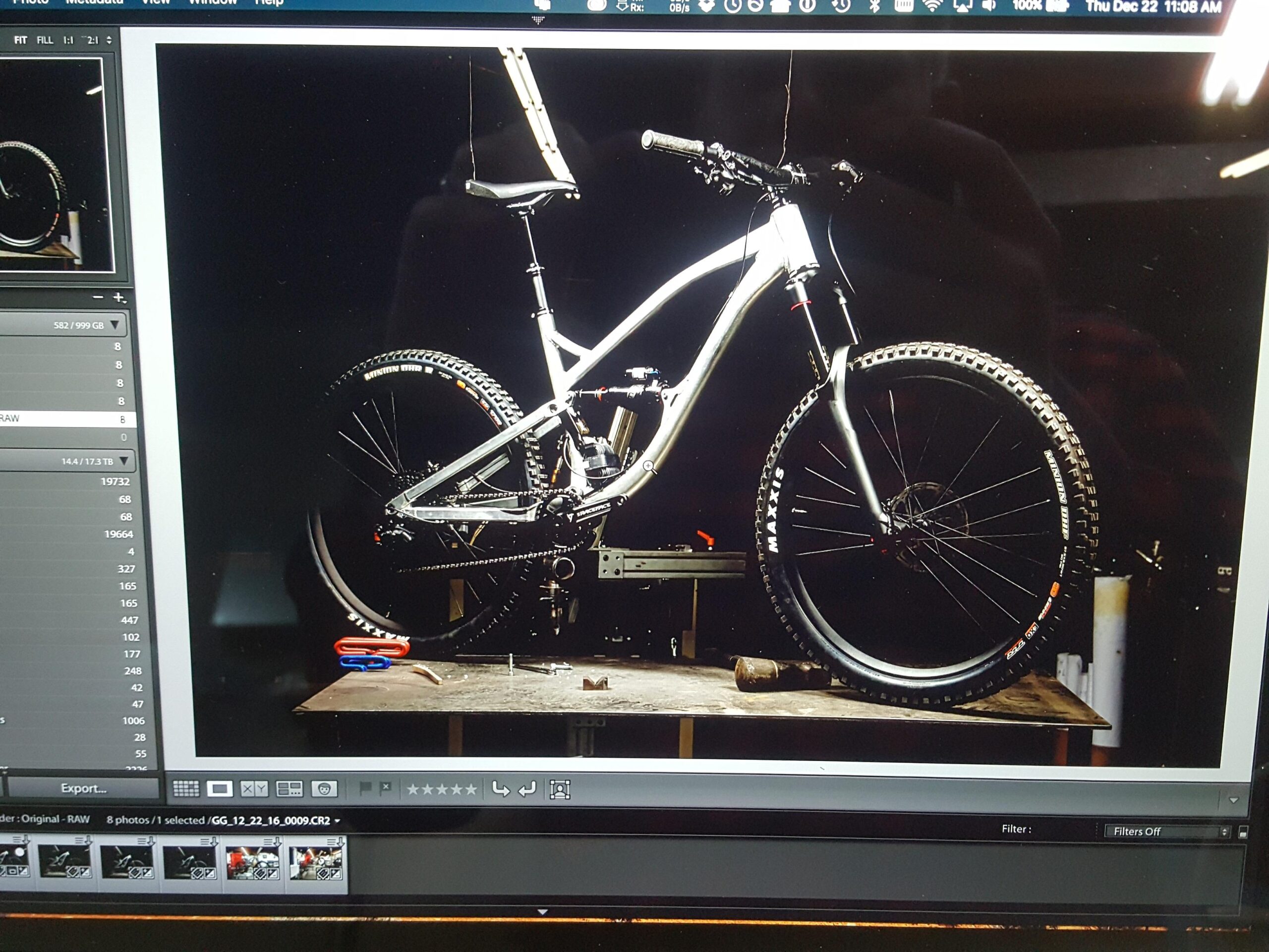 Guerrilla Gravity Megatrail: A mountain bike is displayed on a workbench against a dark background, with tools and accessories scattered nearby. The bike features a shiny silver frame, thick tires, and is in a well-lit, detailed view for inspection or repair.