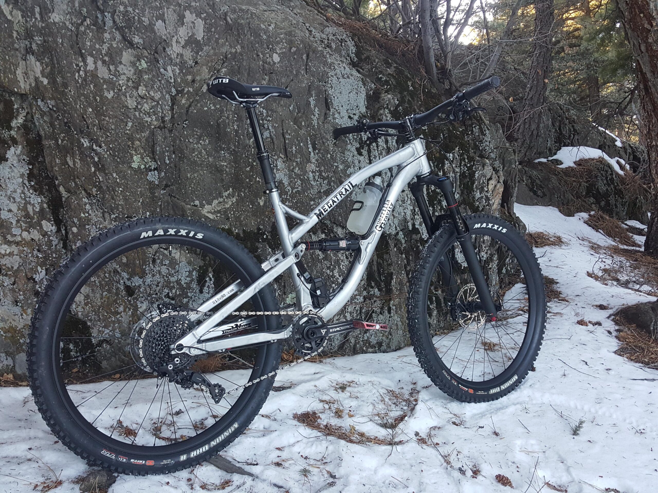 Guerrilla Gravity Megatrail: A silver mountain bike is leaning against a large rock, partially covered in snow and surrounded by trees. The bike features wide tires labeled "Maxxis" and a modern frame design, showcasing its suitability for rugged terrain.
