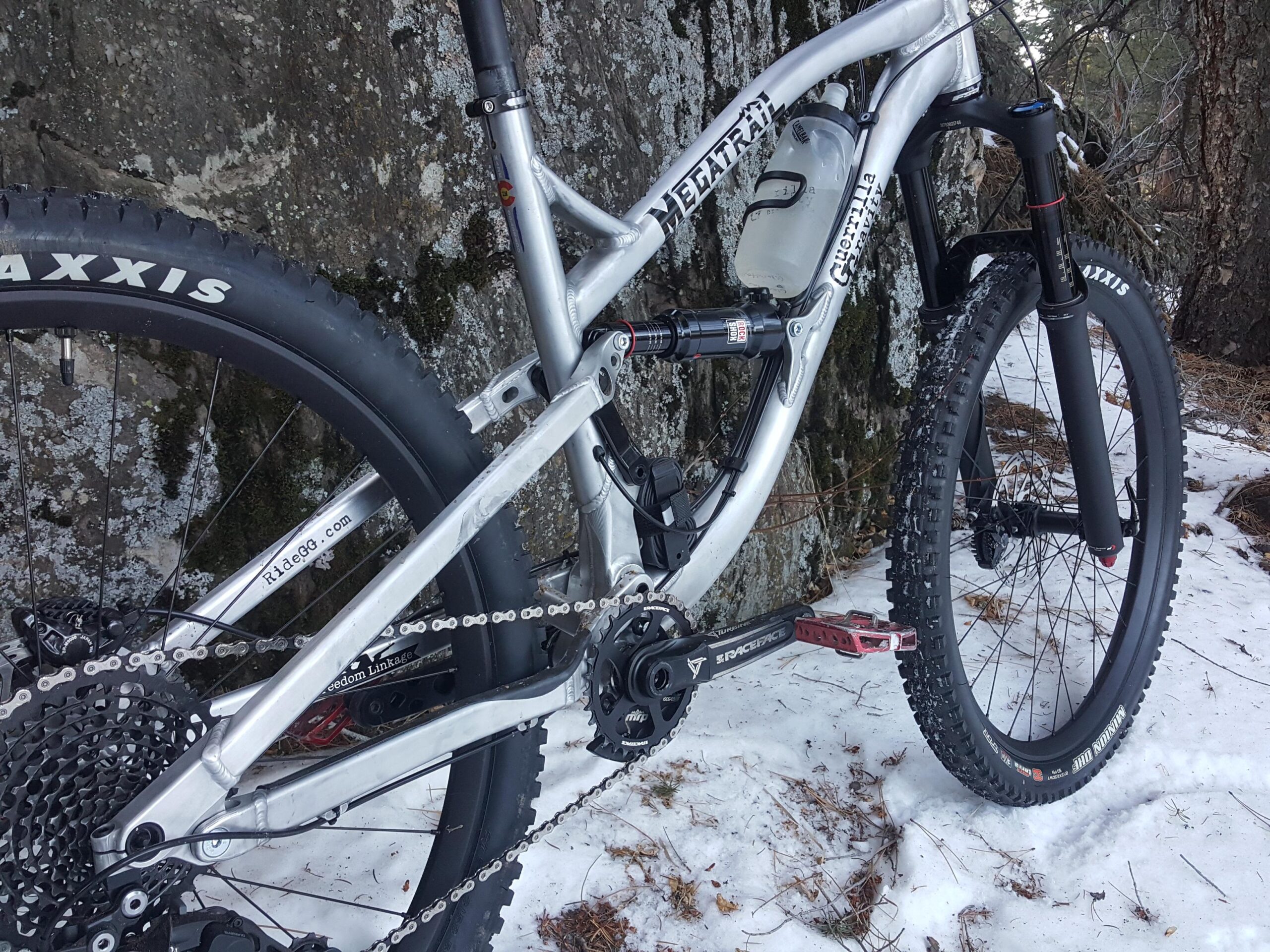 Guerrilla Gravity Megatrail: Close-up view of a silver mountain bike parked on snowy ground next to a tree. The image highlights the bike's frame, wheels, and drivetrain components, including a suspension fork and wide tires with "Maxxis" branding. The background features a rugged natural setting with patches of snow and bark.