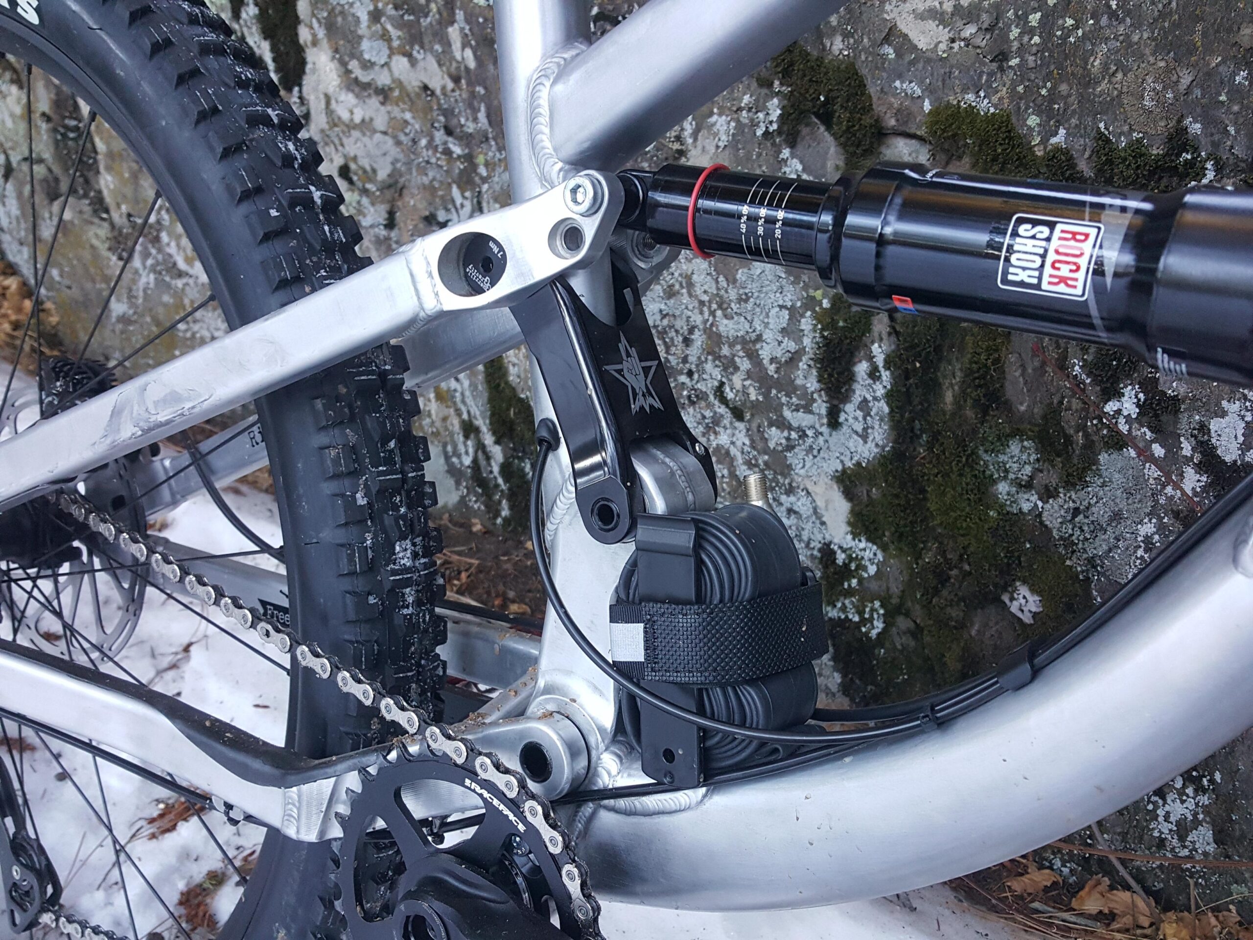 Guerrilla Gravity Megatrail: Close-up view of the rear suspension and drivetrain of a mountain bike, featuring a RockShox shock absorber, chain, and rugged tire. The bike's aluminum frame is shown against a stone surface covered in moss and lichen, with some snow visible on the ground.