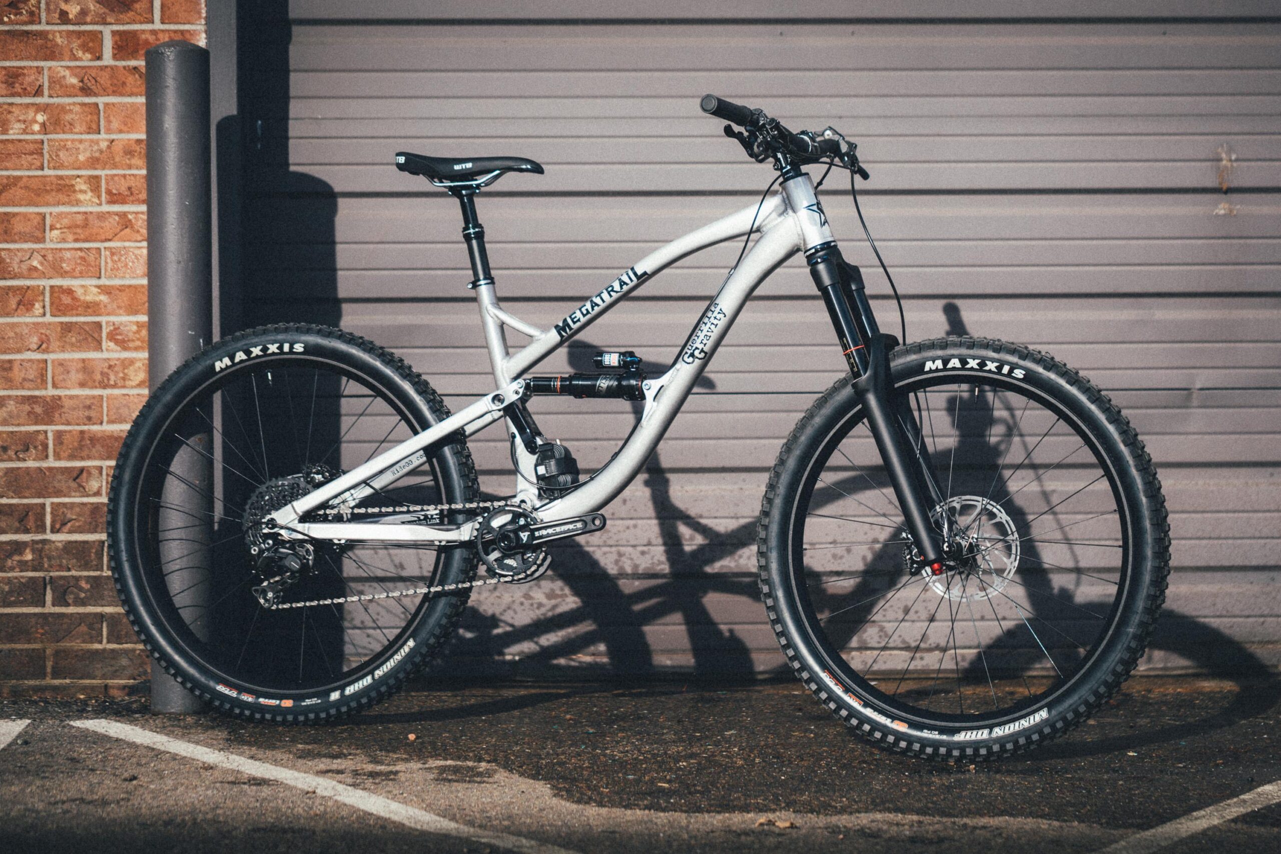 Guerrilla Gravity Megatrail: A silver mountain bike leaning against a textured brick wall, with a black fork and Maxxis tires. The bike features a prominent "Megatrail" logo, showcasing its sturdy frame and components. The background includes a closed garage door.