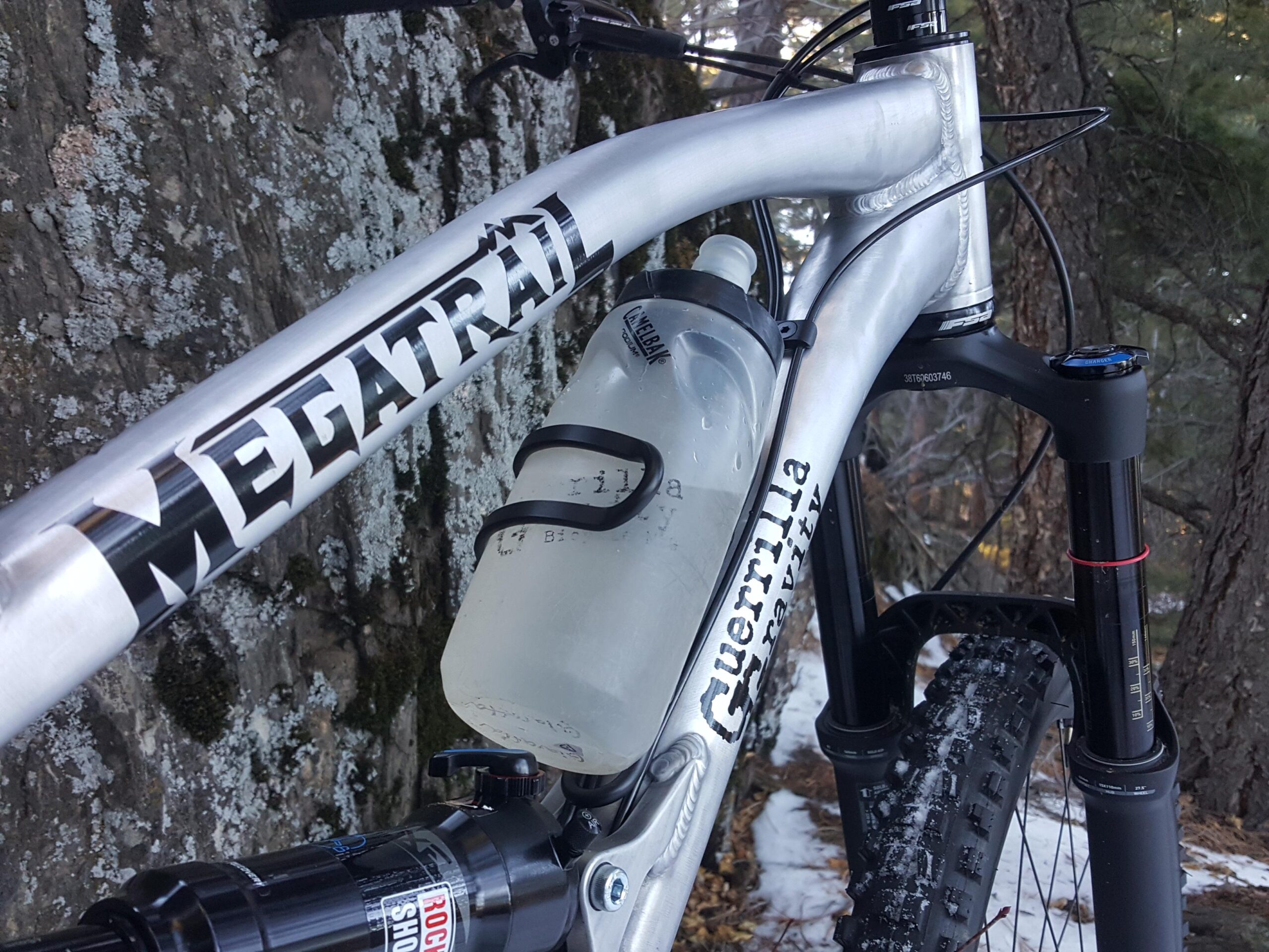 Guerrilla Gravity Megatrail: A close-up view of a mountain bike frame with the words "MEGATRAIL" and "Guerrilla Gravity" visible. The bike features a water bottle mounted in the frame and a rugged tire, set against a backdrop of trees and rocky terrain, indicating a natural outdoor environment. Snow and moss are present, suggesting a cooler climate.