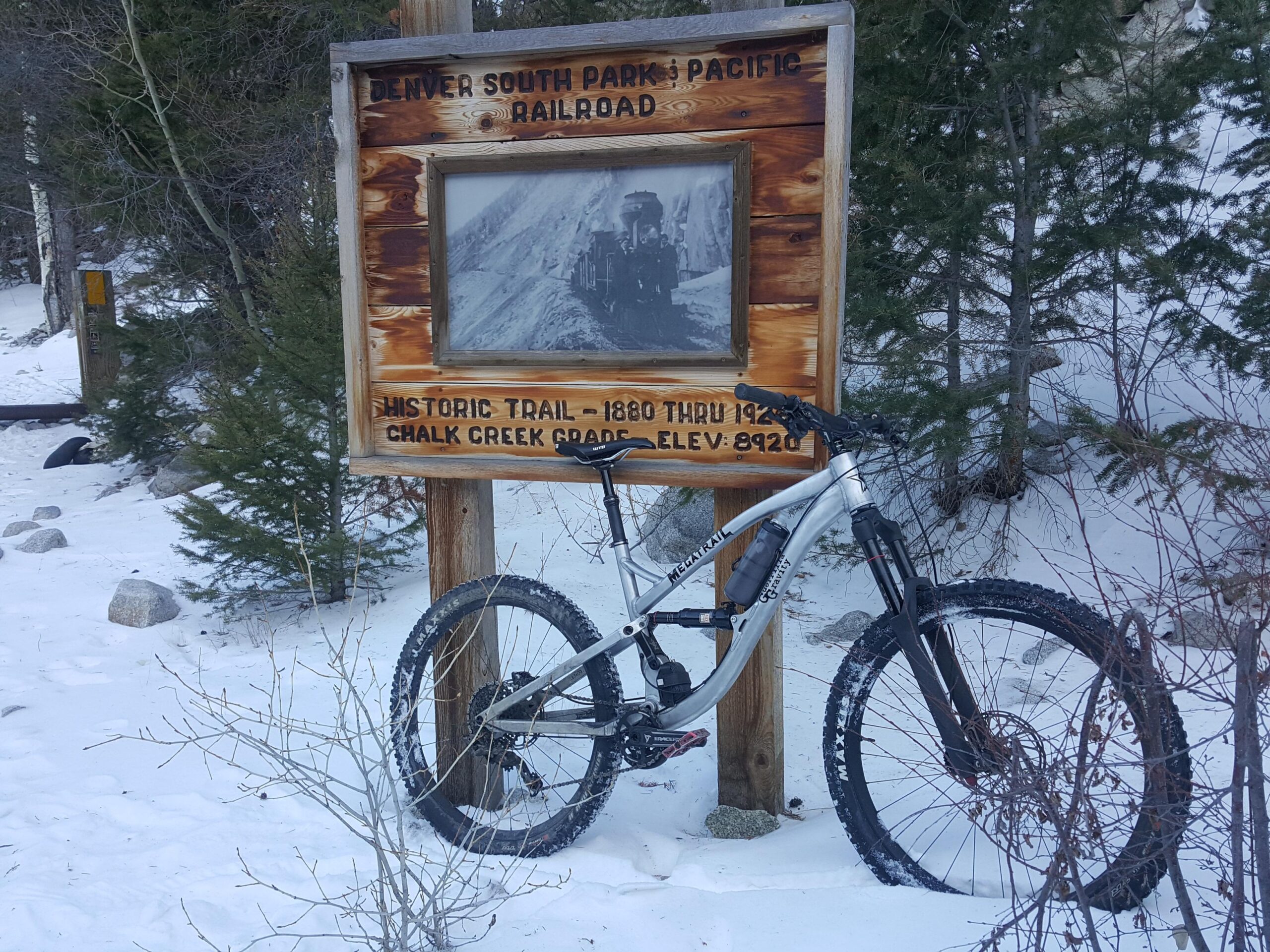 Guerrilla Gravity Megatrail: A mountain bike is leaning against a wooden sign for the Denver South Park & Pacific Railroad, which features historical information about the trail, including its elevation of 8920 feet. The scene is set in a snowy landscape with evergreen trees in the background.