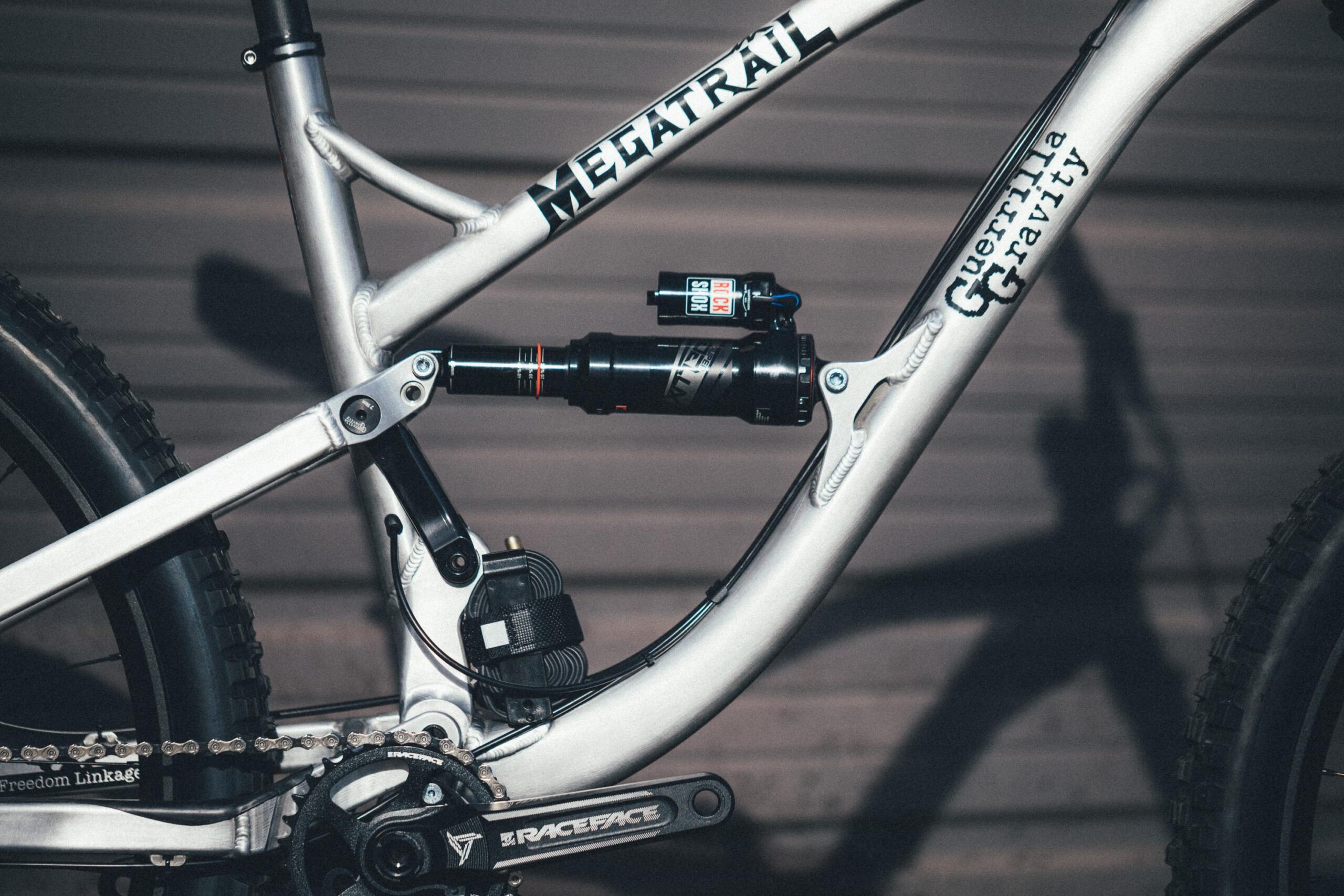Guerrilla Gravity Megatrail: Close-up view of a mountain bike frame showcasing its suspension system, featuring components like the shock absorber, chain, and pedal. The frame is painted in a light color with branding visible, highlighting details of the bike's design and engineering.