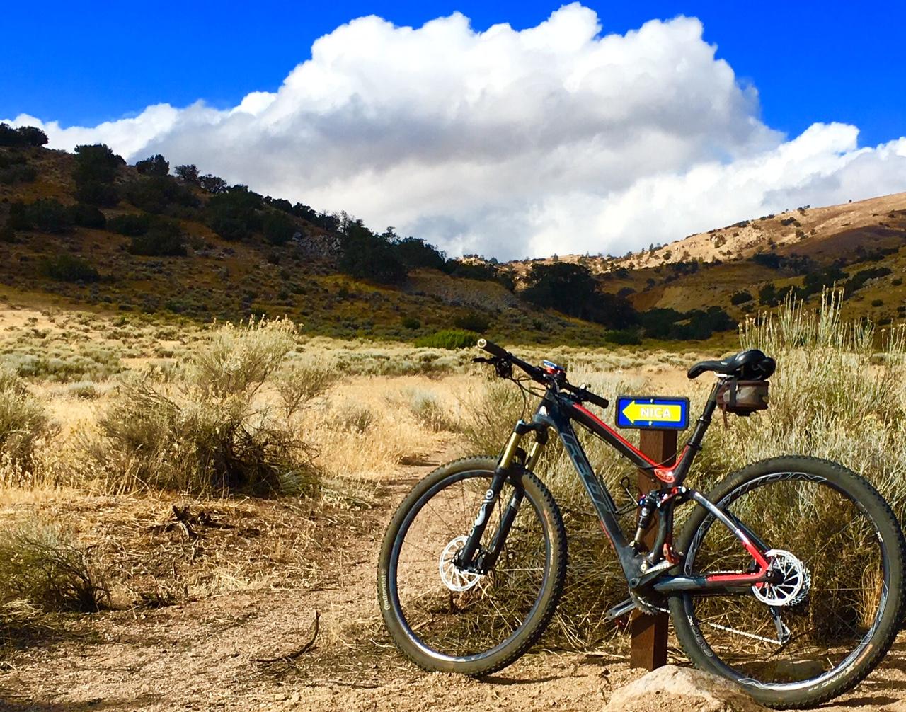 A mountain bike leaning against a post with a directional sign reading "NICA" in a natural landscape, featuring rolling hills, dry grass, and scattered shrubs under a blue sky with fluffy white clouds. TMTA Lehigh trails mountain bike trail.