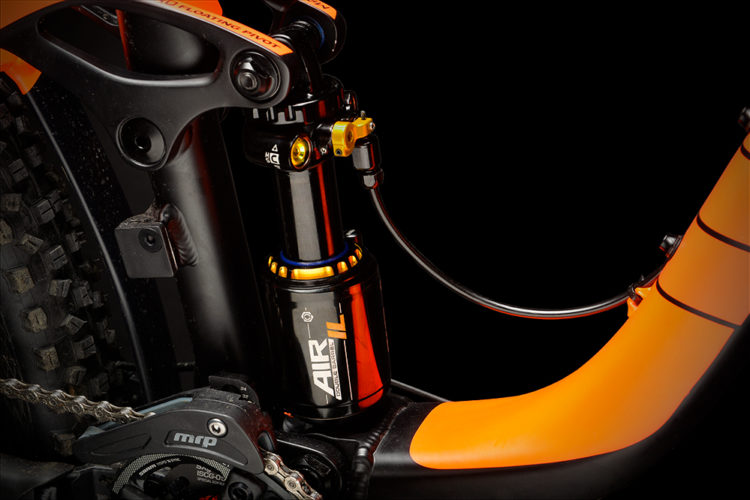 Close-up view of a mountain bike's rear suspension system, featuring a black and orange air shock absorber. The shock is mounted on the bike frame, showcasing detailed components like the linkage and a visible portion of the drivetrain, with a dark background emphasizing the equipment's features.