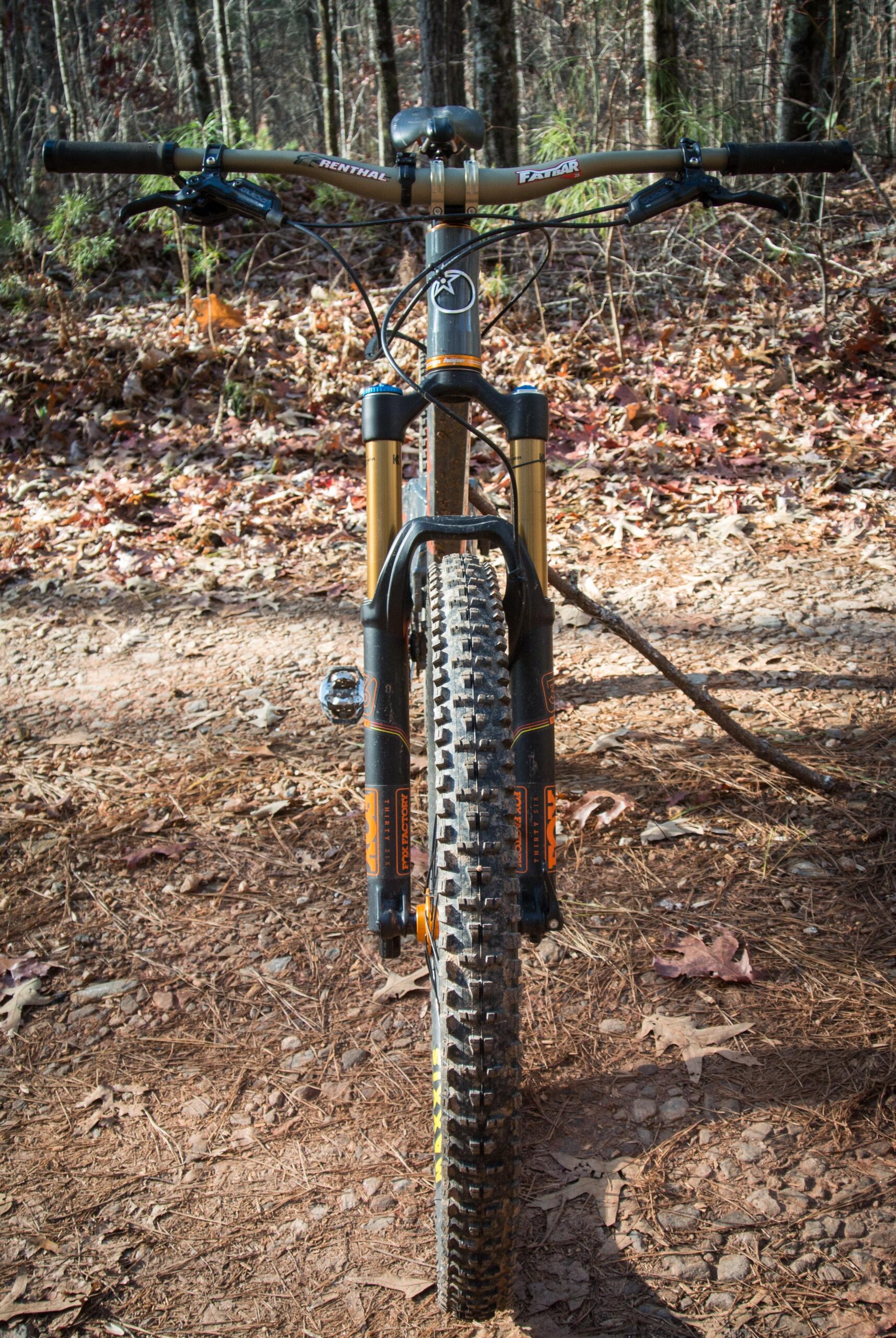 Orange Five RS: A mountain bike viewed from the front, positioned on a dirt trail surrounded by trees. The bike features thick tires, a sleek frame, and suspension forks with gold accents. Fallen leaves and pine needles cover the ground, indicating an autumn setting.