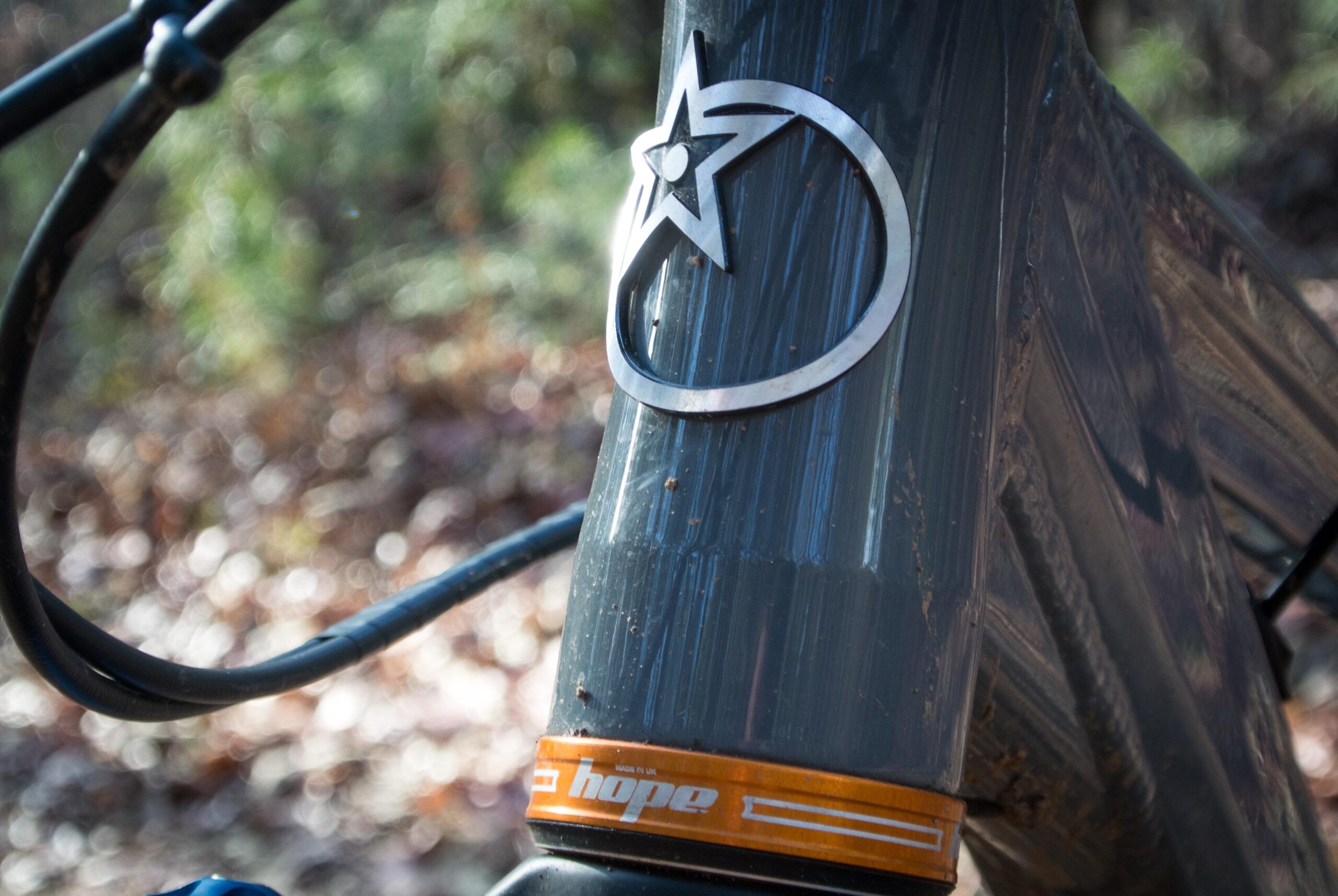 Orange Five RS: A close-up of a mountain bike's head tube featuring a metallic star logo and an orange "hope" brand label. The background shows blurred natural foliage, suggesting an outdoor setting. Dirt and scratches on the bike indicate recent use.