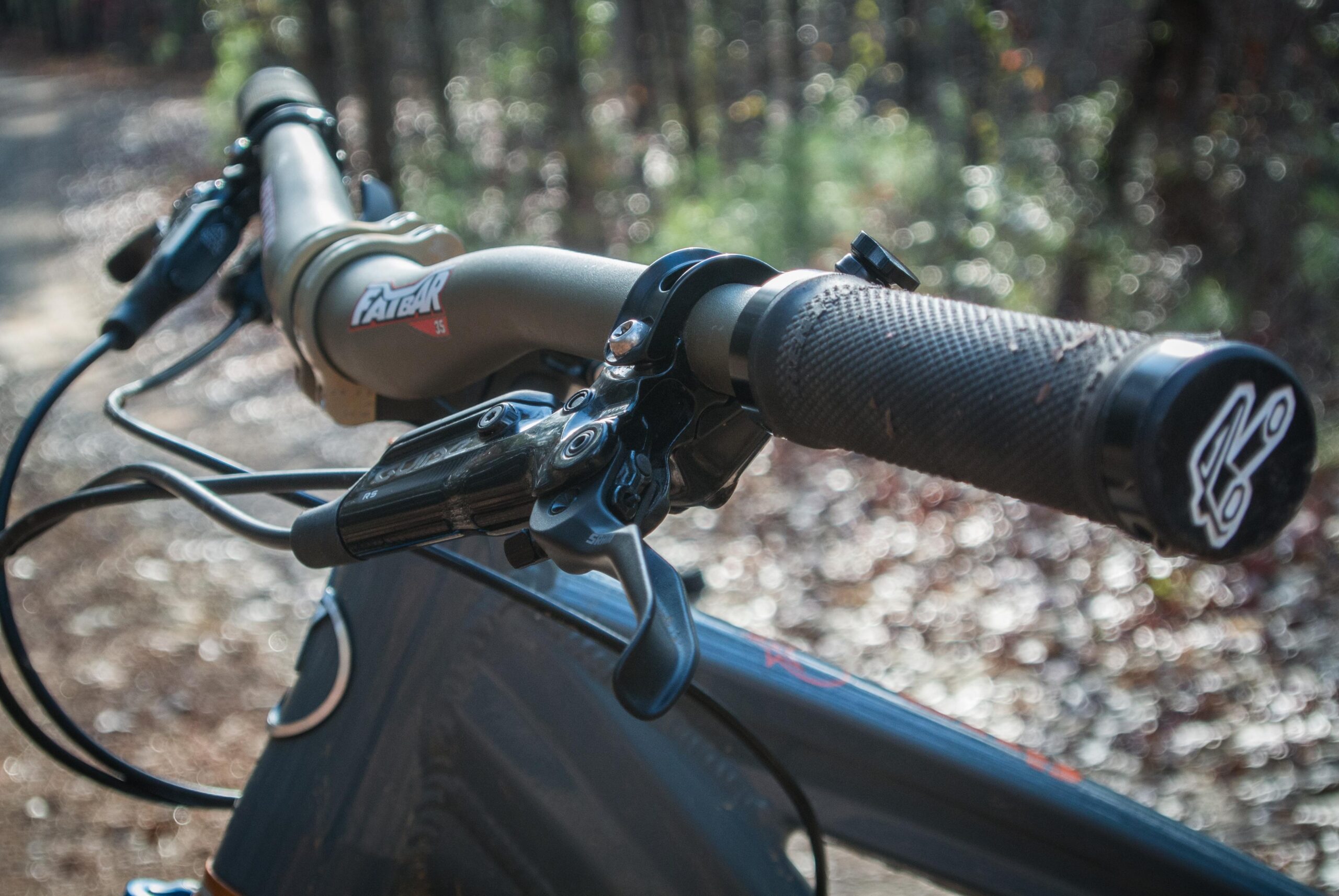 Orange Five RS: Close-up view of a mountain bike's handlebars, featuring grips, brake levers, and a stem with branding. The background shows a blurred forest path with natural lighting.