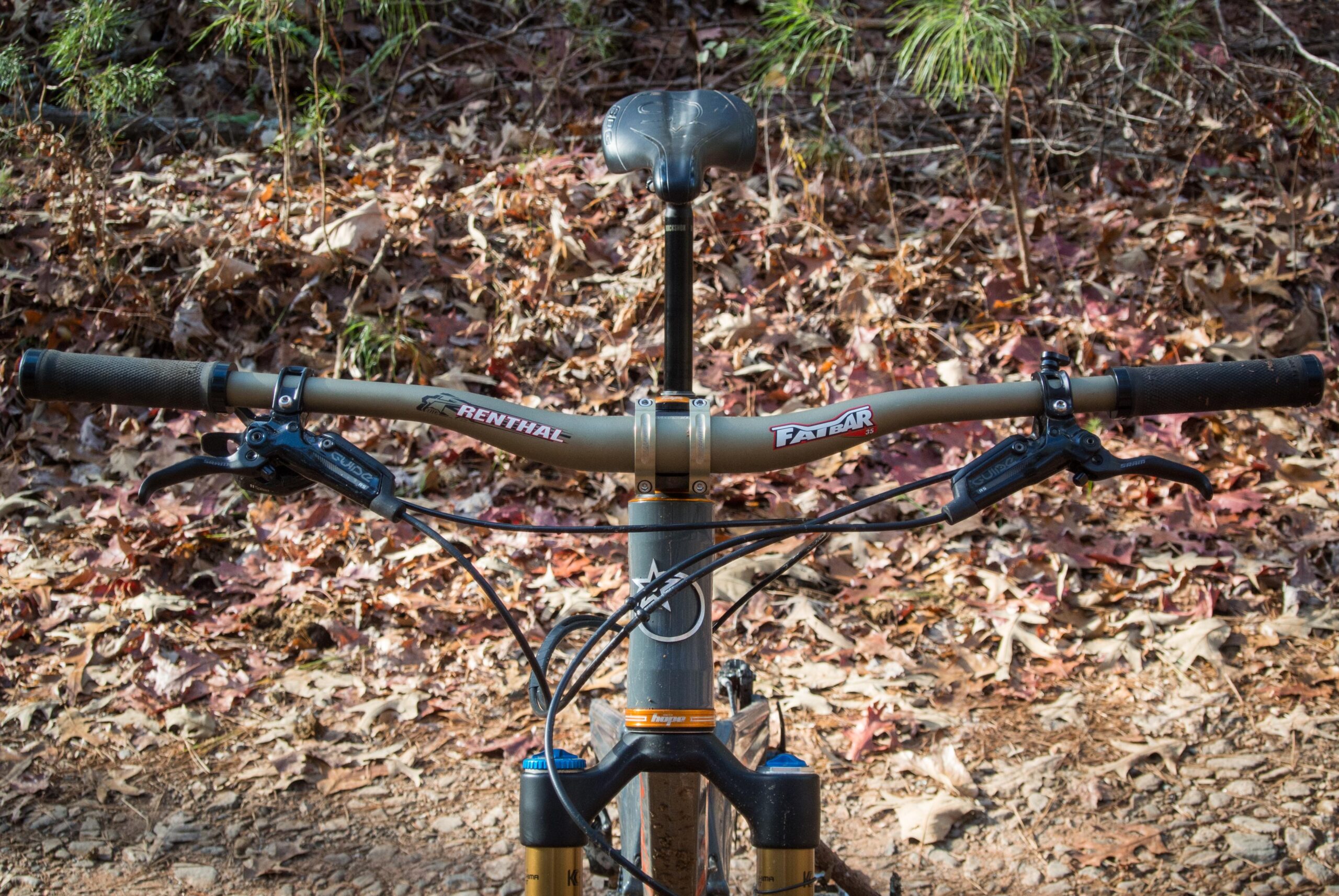 Orange Five RS: Close-up view of a mountain bike's handlebars, featuring Renthal and Fatbar grips, brake levers, and cables, set against a background of fallen leaves and a dirt path in a natural outdoor environment.