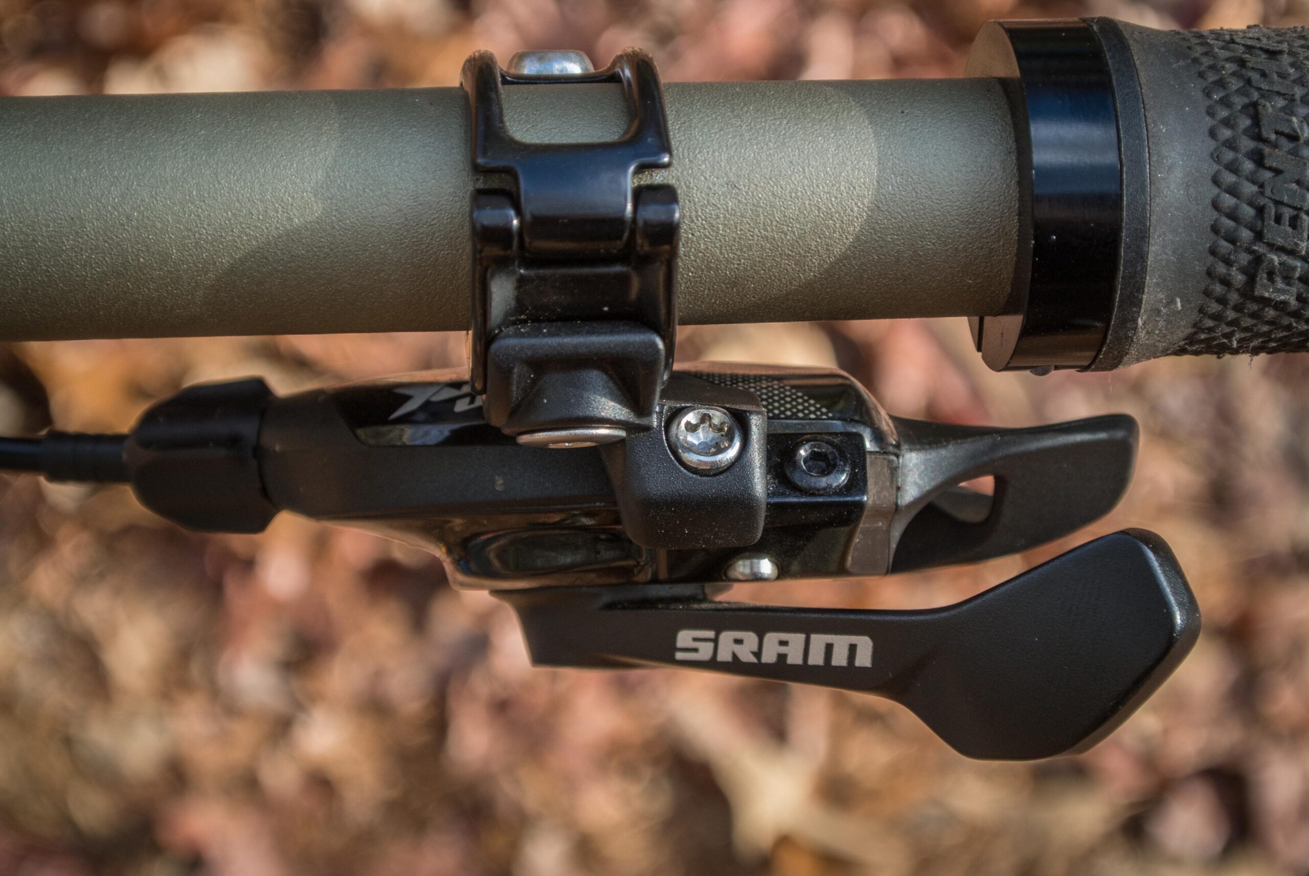 Orange Five RS: Close-up image of a bike handlebar featuring a SRAM gear shifter and a textured grip. The handlebar is olive green with a black clamp attachment. The background consists of blurred autumn foliage.