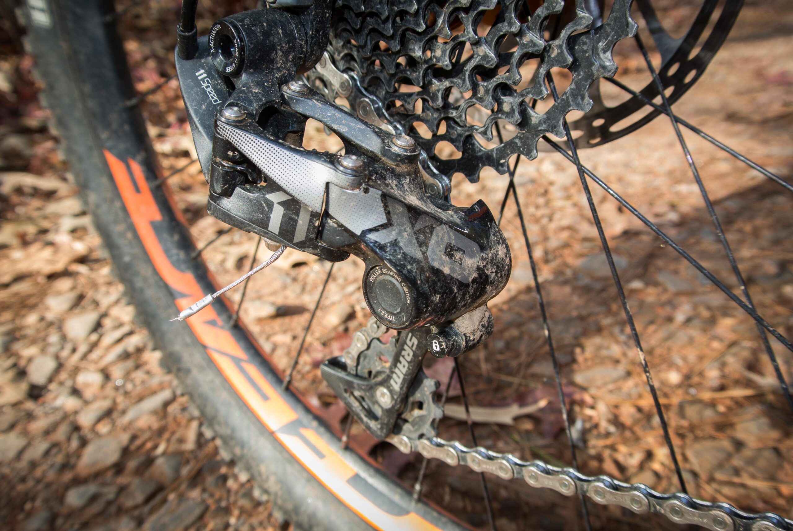 Orange Five RS: Close-up of a bicycle's rear derailleur, featuring a mix of black and silver components with some dirt and debris. The derailleur is attached to a chain and sits near a bike wheel with an orange stripe. The background shows a textured surface of rocks and leaves.