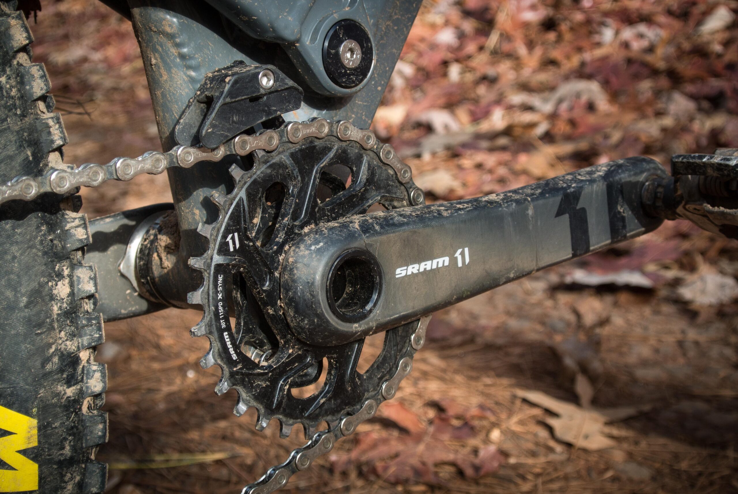 Orange Five RS: Close-up of a bicycle's crankset and chain, featuring a SRAM component. The crank and chainrings are slightly covered in dirt, set against a backdrop of pine needles and scattered leaves.