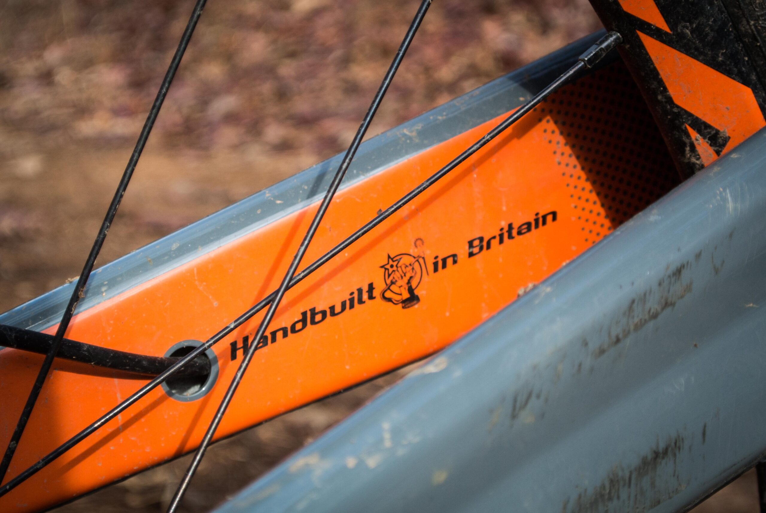 Orange Five RS: Close-up of a bicycle frame highlighting an orange section with the text "Handbuilt in Britain" and a logo. The frame shows signs of use with dirt and scratches.