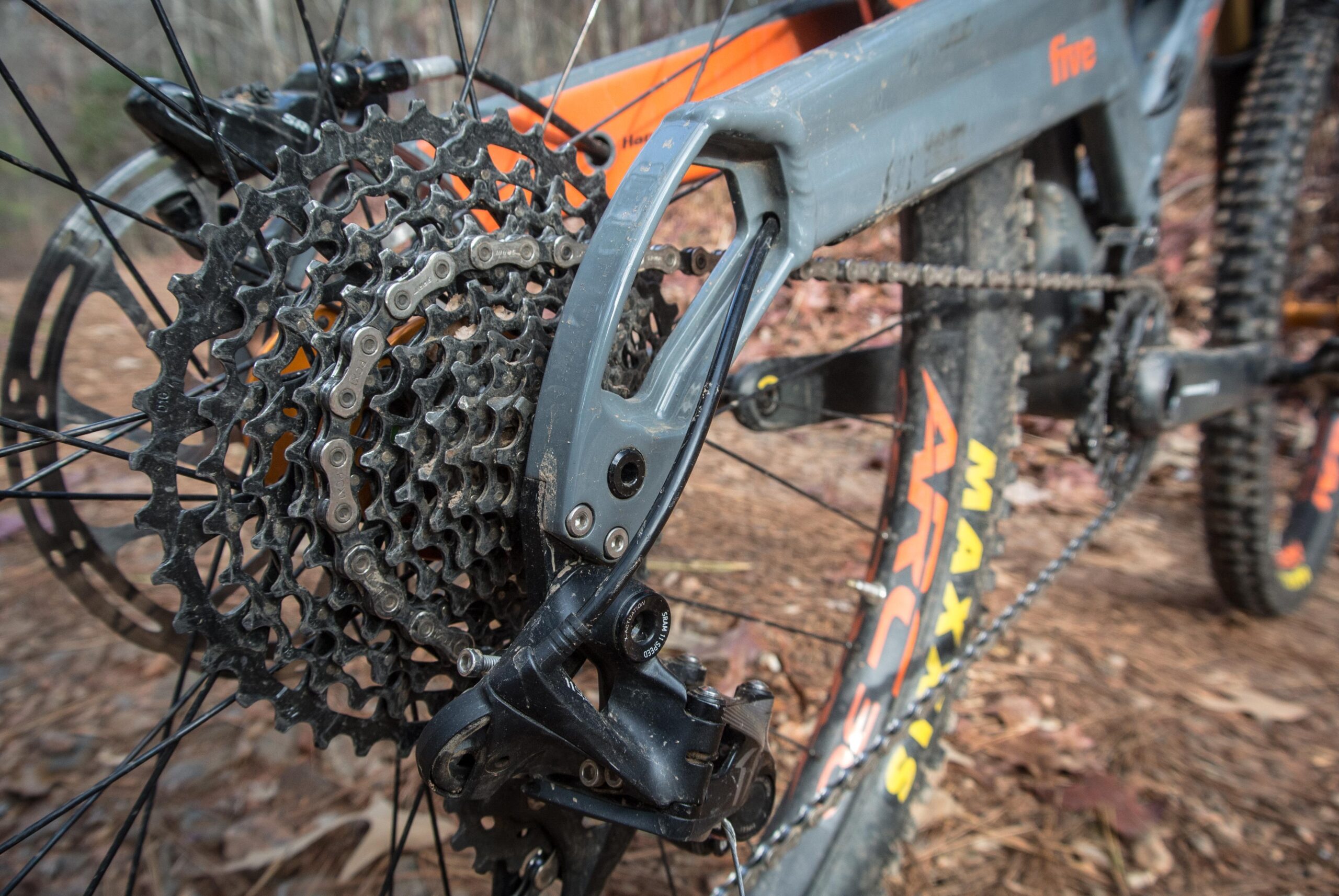 Orange Five RS: Close-up view of a mountain bike's rear cassette and derailleur, showcasing the intricate gear details and chain components. The bike frame is partially visible, featuring orange accents against a gray finish, with a natural, earthy background.
