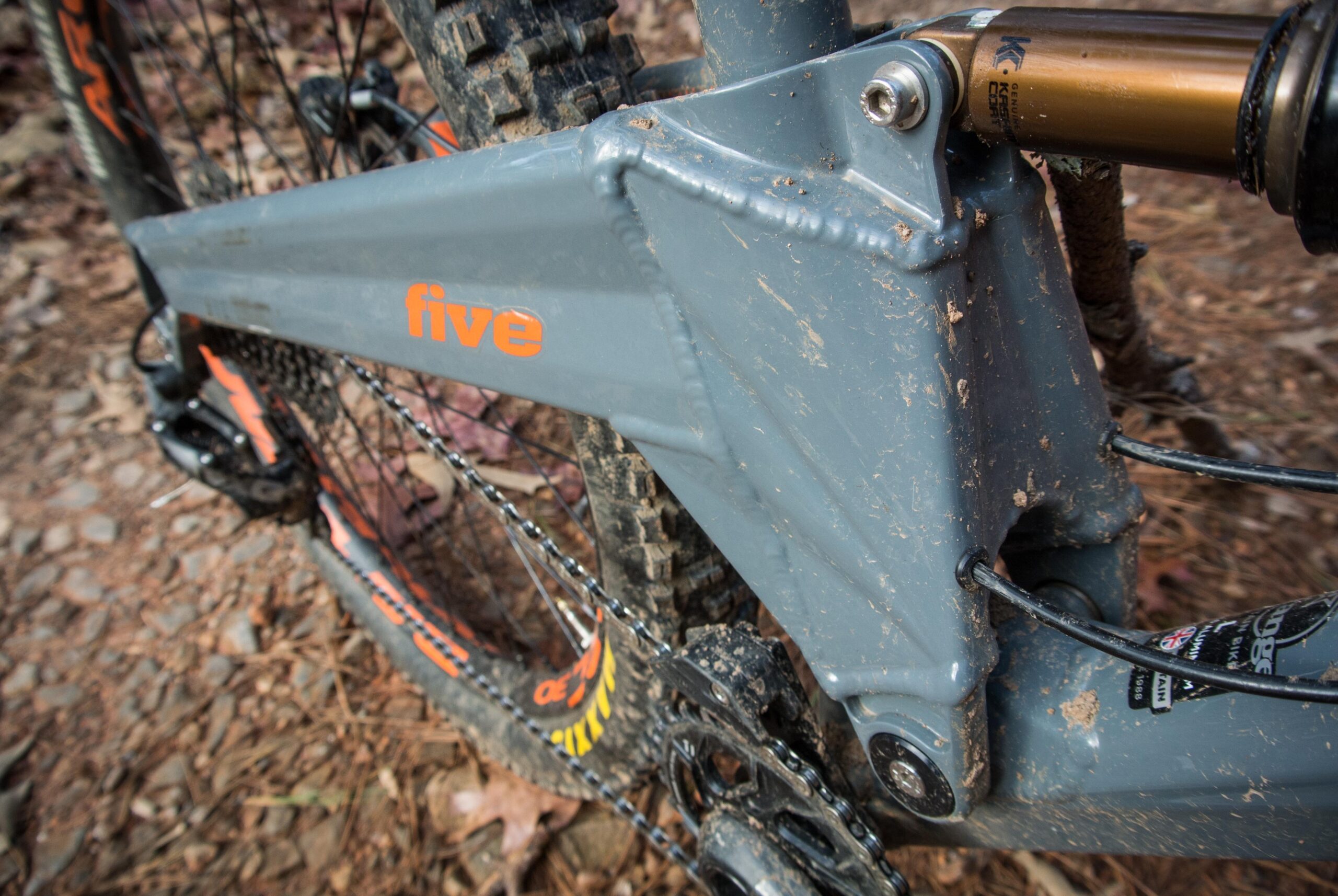 Orange Five RS: Close-up view of a mountain bike frame showcasing the suspension linkage and chainstay. The frame is gray with an orange logo reading "five," and it's partially covered in dirt, indicating recent use on a trail. The background features leaves and gravel, reflecting an outdoor setting.