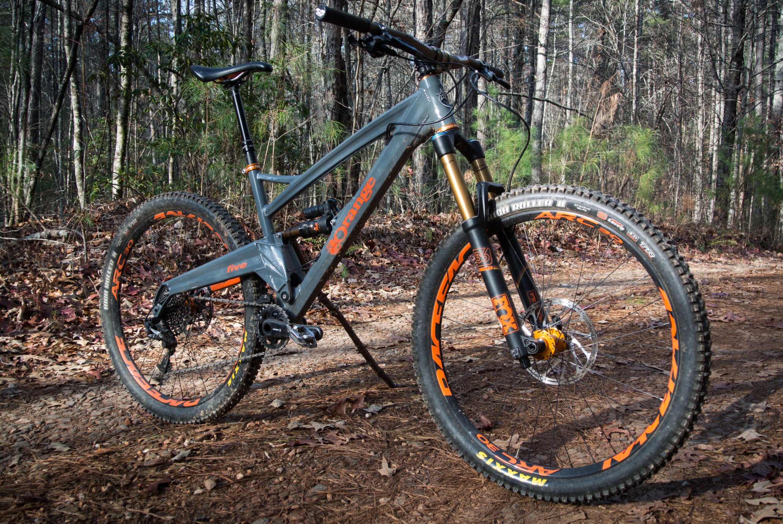 Orange Five RS: A mountain bike with a gray frame and orange accents is positioned on a dirt trail surrounded by trees and fallen leaves. The bike features large, knobby tires and a sturdy fork, indicating its suitability for off-road terrain.