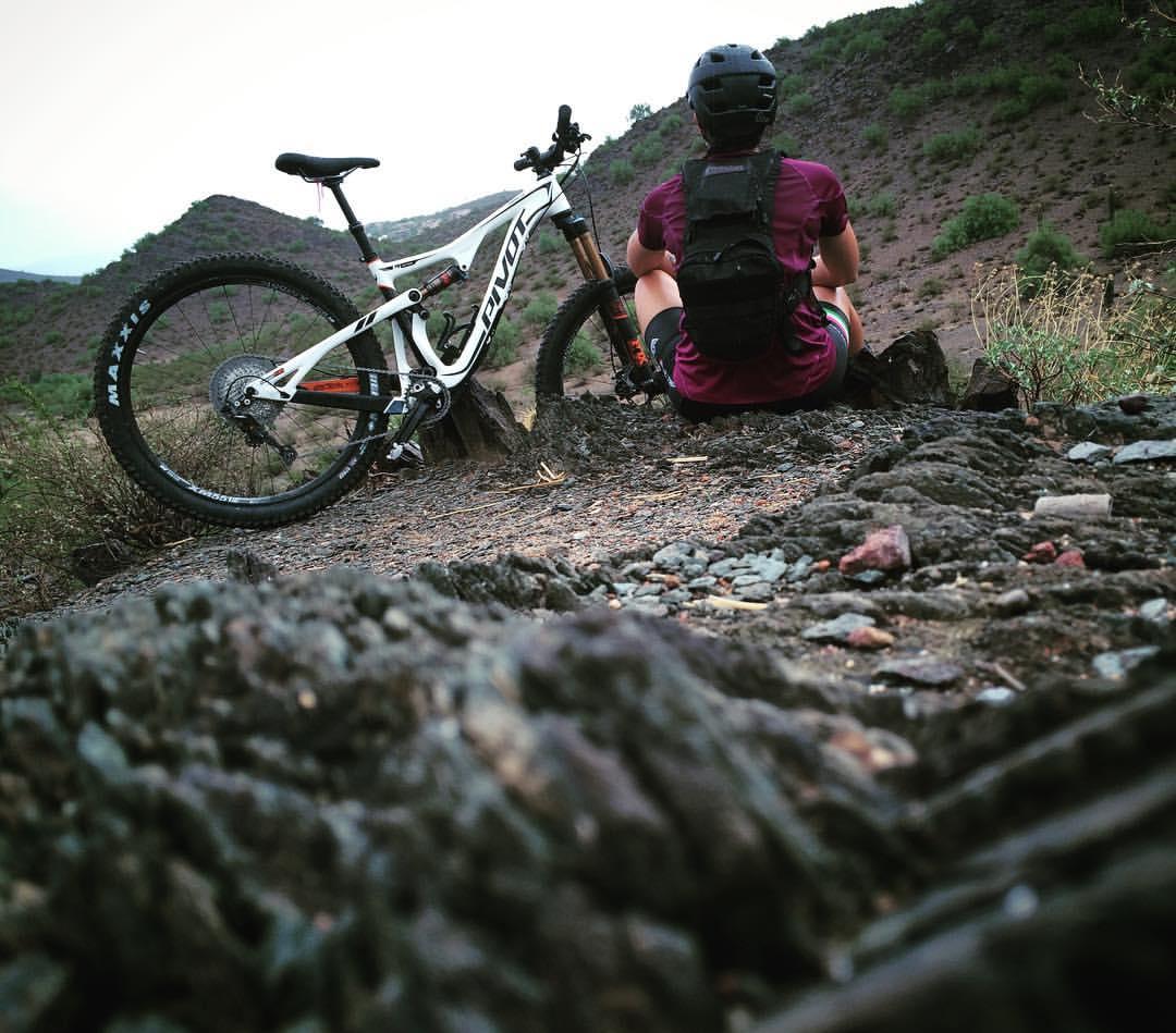 Pivot Trail 429: A person sitting on a rocky trail, looking out towards a mountainous landscape, with a mountain bike leaning beside them. The scene captures a sense of adventure and contemplation in a natural setting.