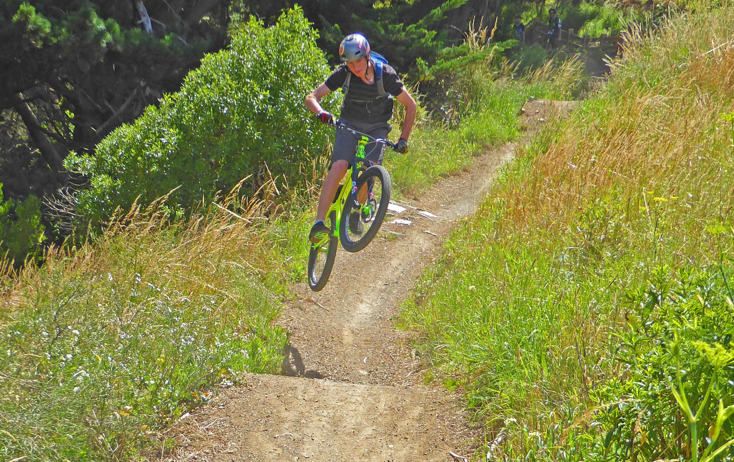 A cyclist performing a jump on a dirt trail surrounded by greenery and tall grass on a sunny day. The rider is wearing a helmet and gloves, showcasing a dynamic biking moment. Mt Victoria mountain bike trail.