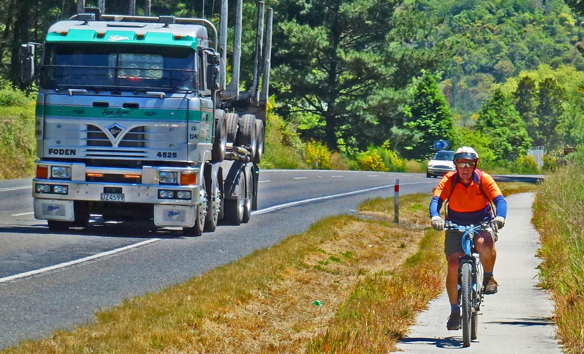 A cyclist wearing an orange shirt and helmet rides on a path next to a busy roadway, alongside a large green and silver truck traveling in the opposite direction. Lush trees and greenery line the sides of the road in a sunny outdoor setting. Go Thermal by Bike - Te Ara Ahi mountain bike trail.