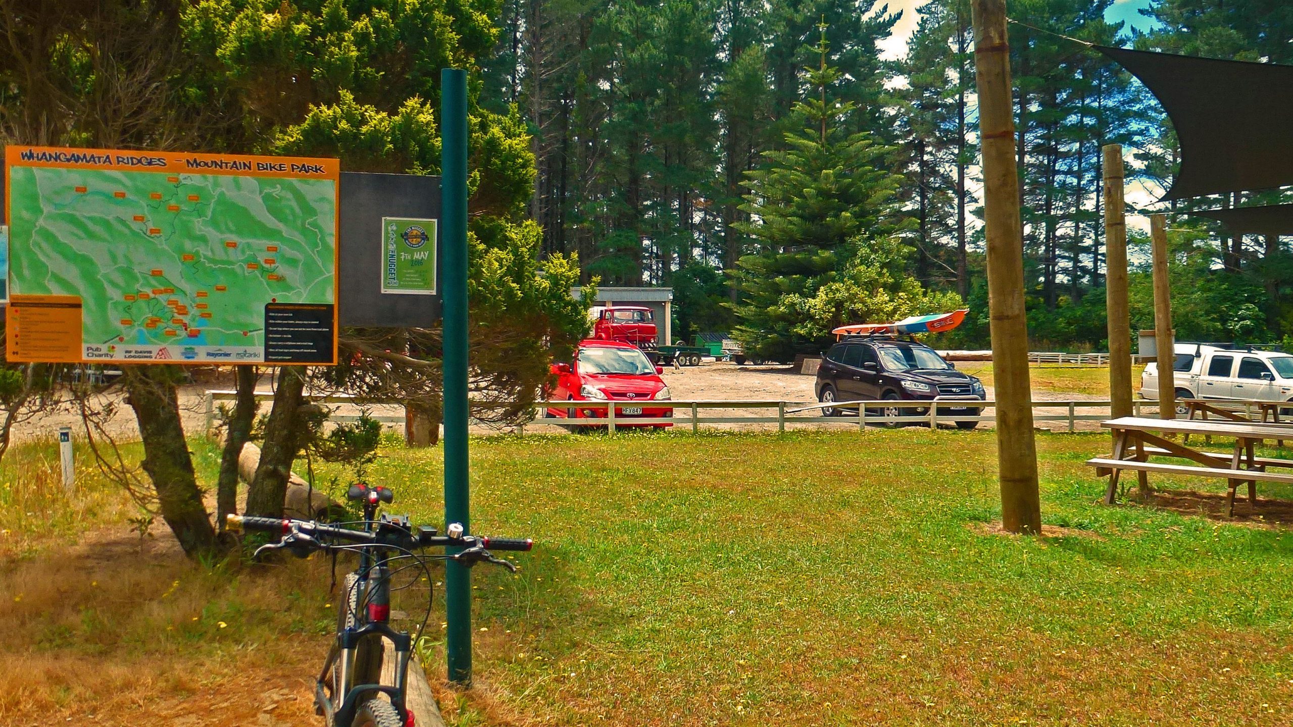 A sign with a map of the Whangamata Ridges Mountain Bike Park is displayed prominently in a grassy area. In the background, various parked vehicles, including a red car and a kayak on top of an SUV, are visible, along with trees and a small building. A bicycle is leaning against a tree on the left side of the image. Whangamata Ridges Mountainbike Park mountain bike trail.