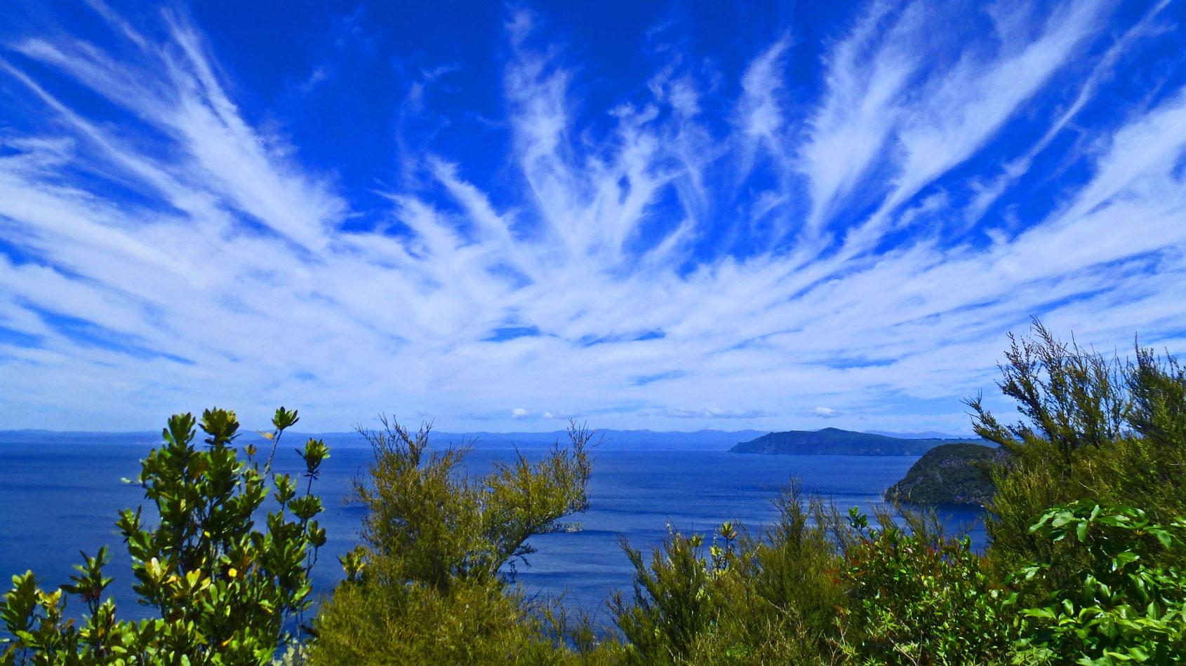 A panoramic view of a serene ocean landscape, featuring a vibrant blue sky streaked with wispy white clouds. In the foreground, lush green foliage frames the scene, while distant mountains are visible across the water. The tranquil blue sea reflects the clear sky, creating a peaceful atmosphere. Great Lake Trail - W2K mountain bike trail.