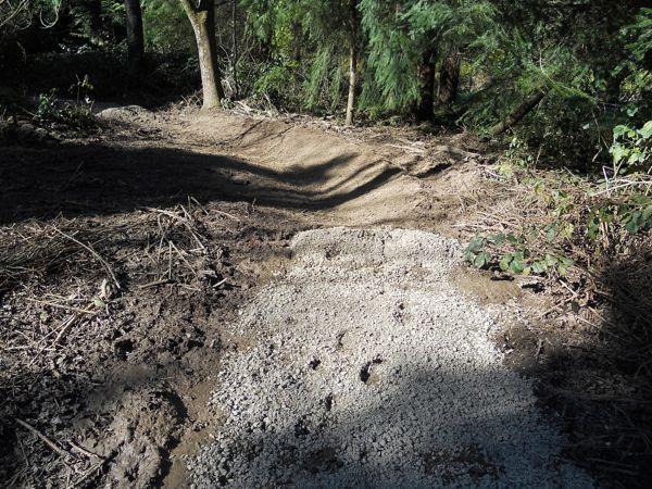 A partially cleared forest path with exposed soil and gravel, surrounded by trees and underbrush. Sunlight casts shadows on the uneven terrain, indicating recent construction or maintenance work. Raincliff mountain bike trail.