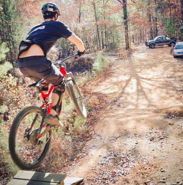 A mountain biker performing a jump off a small ramp on a dirt trail surrounded by trees, with parked cars visible in the background. The scene is set on a sunny day with autumn foliage. Chewacla State Park mountain bike trail.