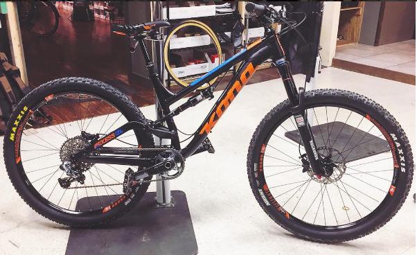 Kona Process 134 Supreme: A black mountain bike with orange and blue accents displayed on a stand in a bike shop. The bike features thick tires, a sturdy frame, and a gear system, showcasing its design for off-road cycling. Background elements include bike accessories and a wooden floor.