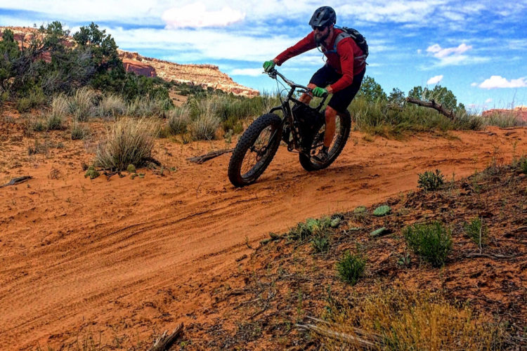 A person riding a fat tire bike on a sandy trail surrounded by desert vegetation and rocky formations, under a partly cloudy blue sky.
